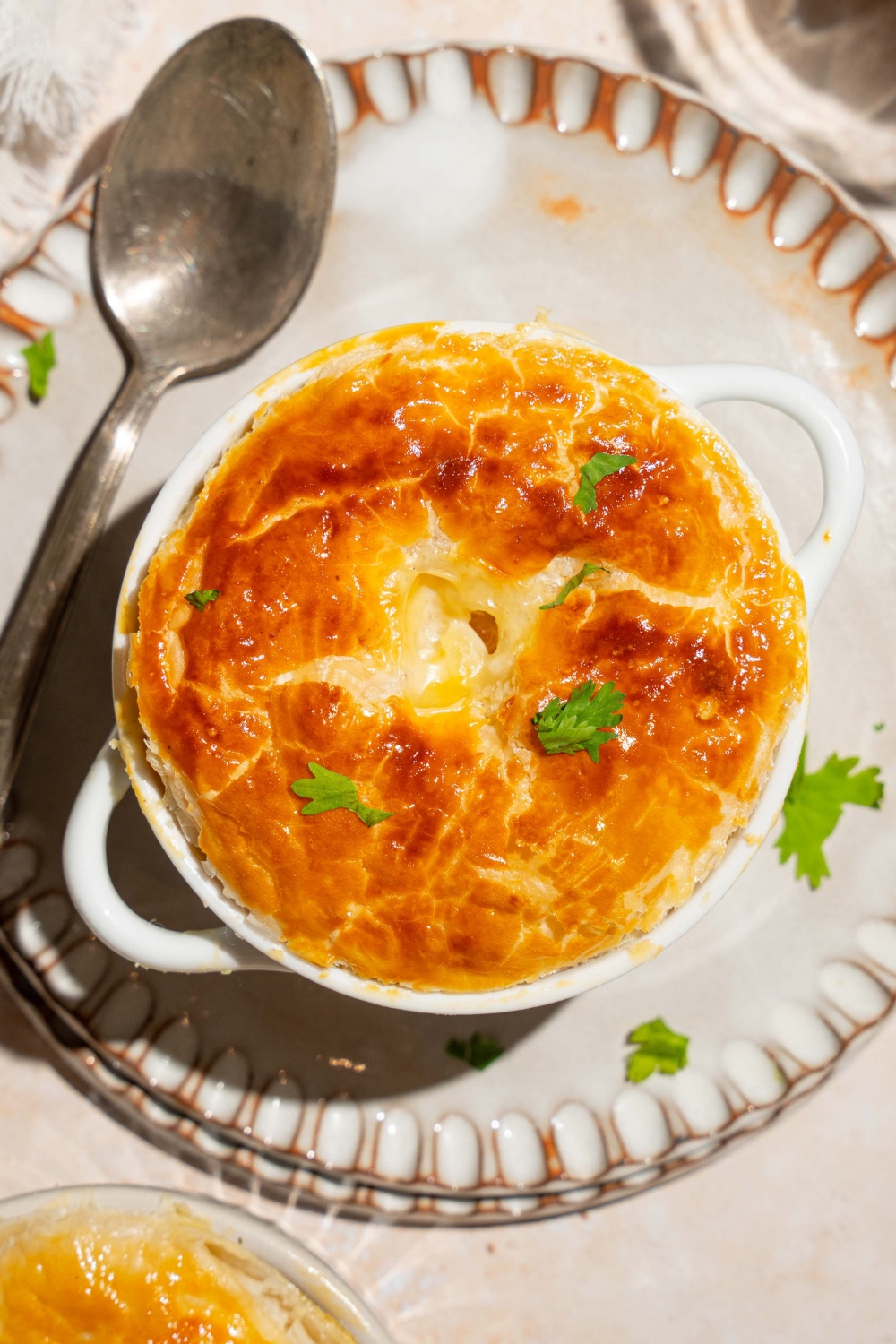 A small pie tin with baked KFC pot pie topped with puff pastry and garnished with fresh parsley. The tin is on plate with a spoon. The plate is on a tan counter.