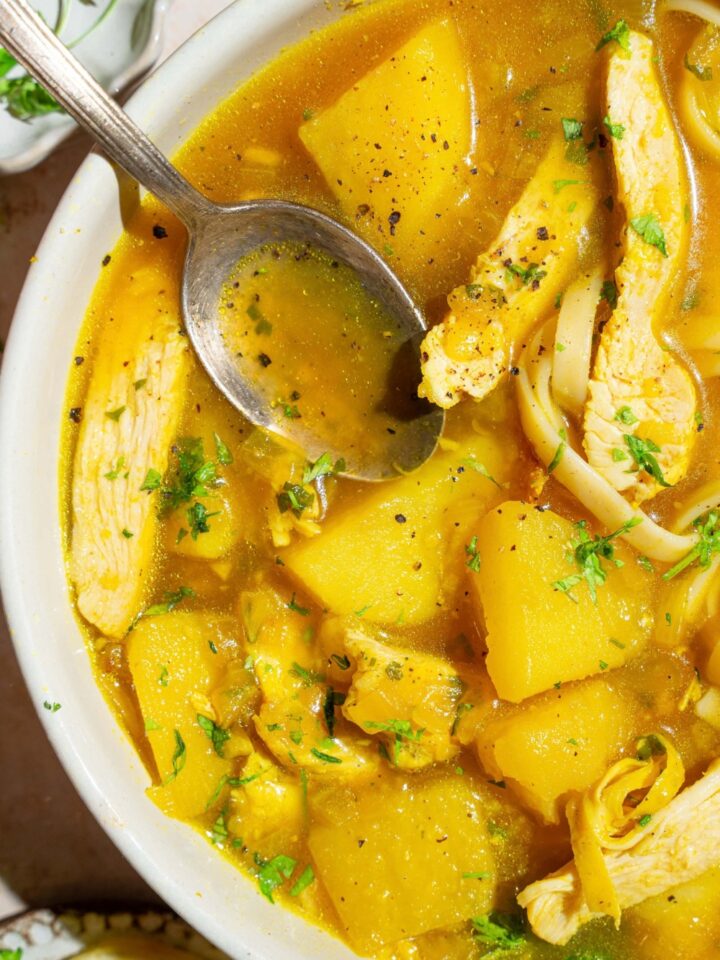 A large crock with garlic chicken soup with fresh parsley. There is a spoon in the pot. The pot is on a tan counter.