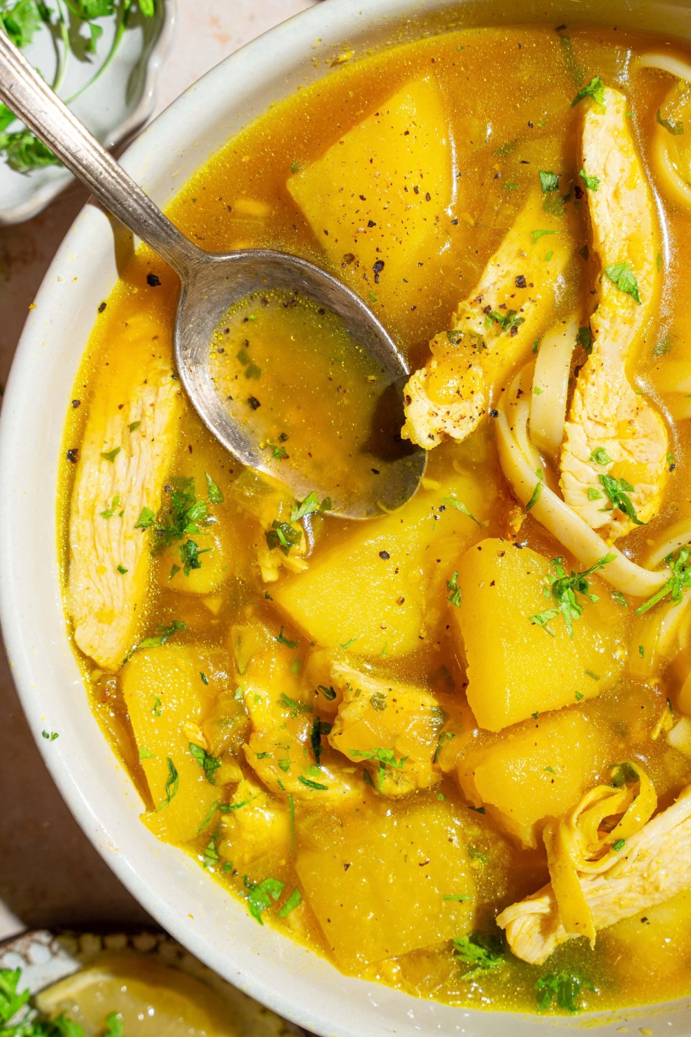 A large crock with garlic chicken soup with fresh parsley. There is a spoon in the pot. The pot is on a tan counter.