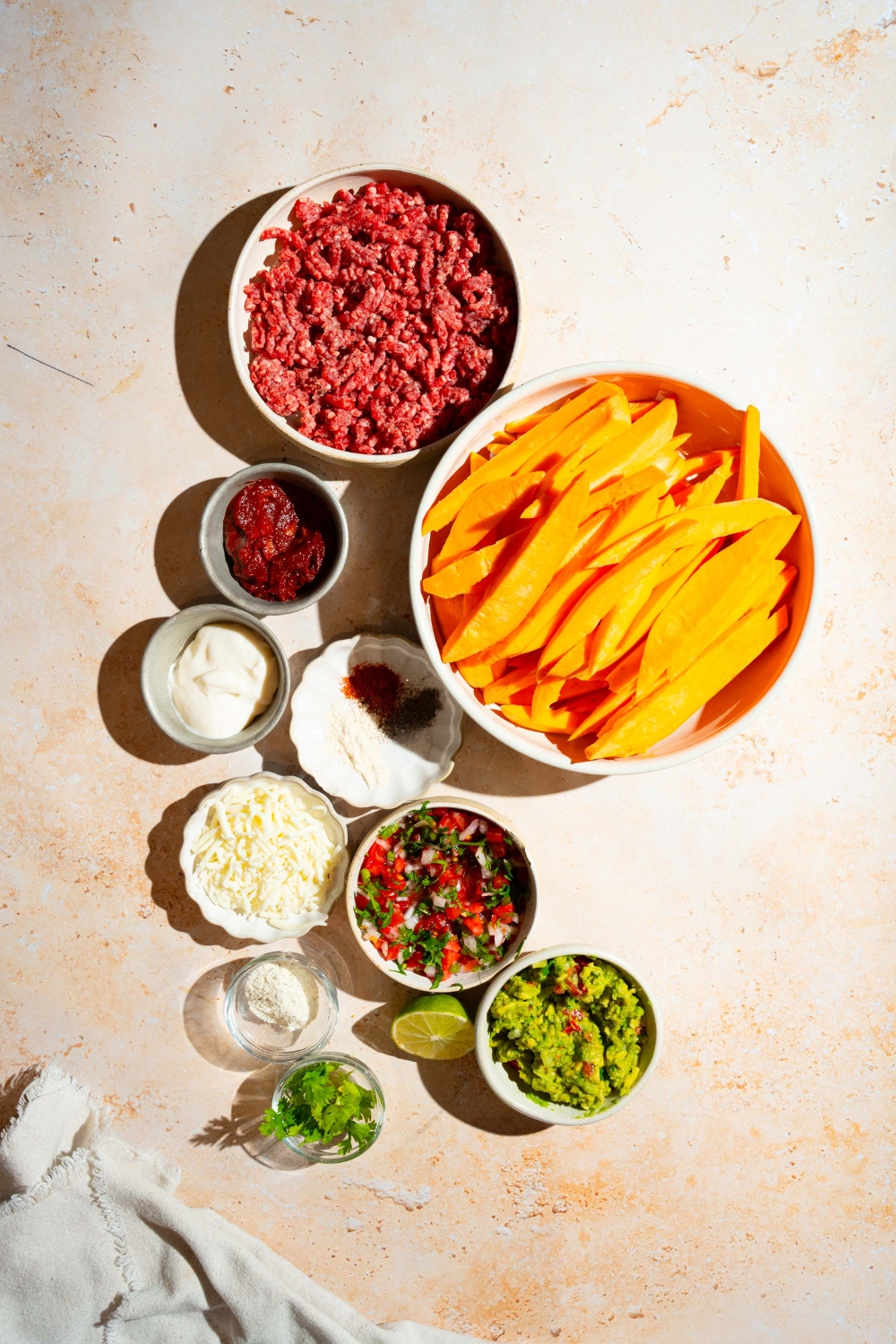 A tan counter with several bowls containing ingredients to make loaded sweet potato fries including sweet potato fries, ground beef, pico de gallo, guacamole, shredded cheese, greek yogurt, lime, and seasonings.