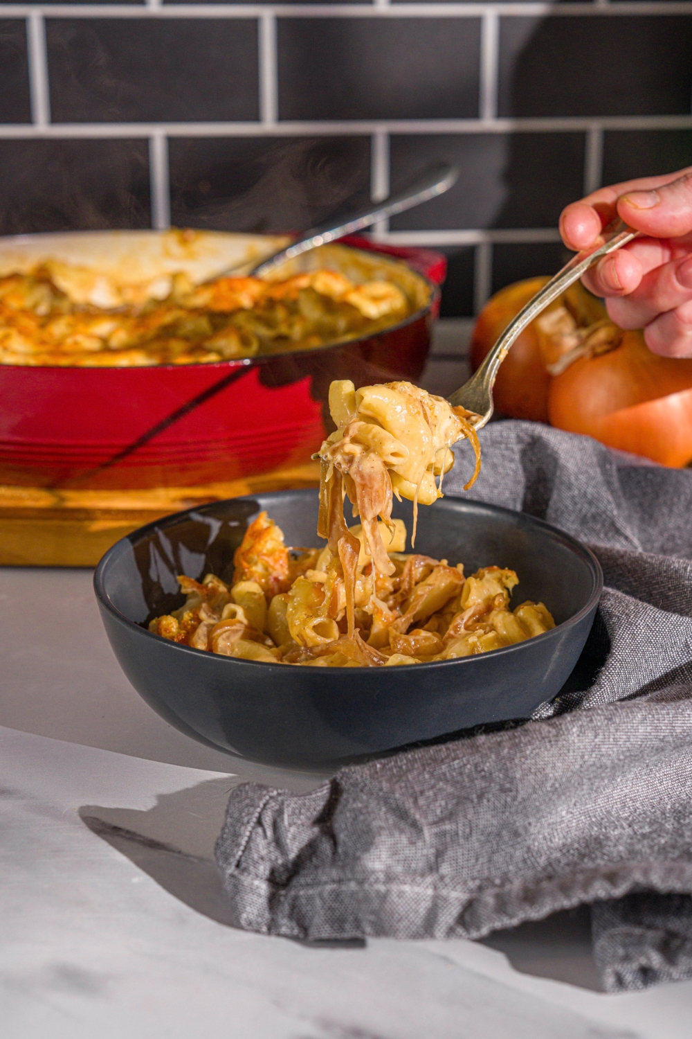 A bowl of French onion mac and cheese. A fork is taking a bite from the bowl. The bowl is on a marble counter with a blue cloth napkin and skillet of mac and cheese.