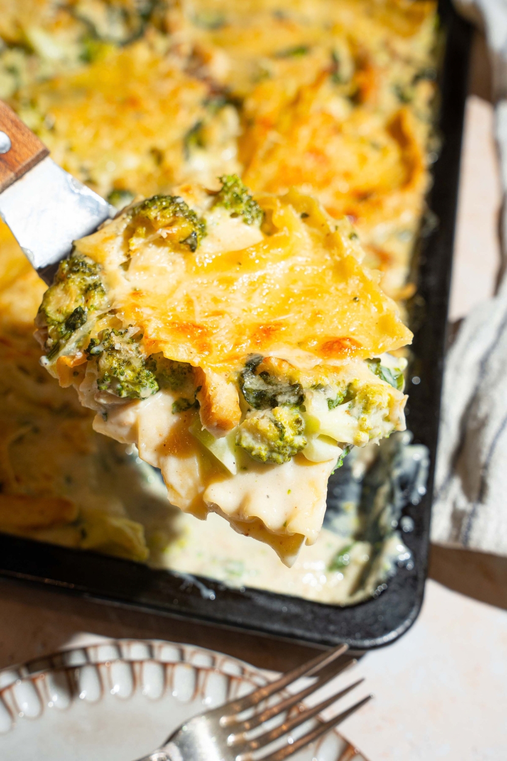 A close up of a spatula with a slice of chicken and broccoli lasagna over a sheet pan of lasagna. The sheet pan is on a tan counter with a plate.