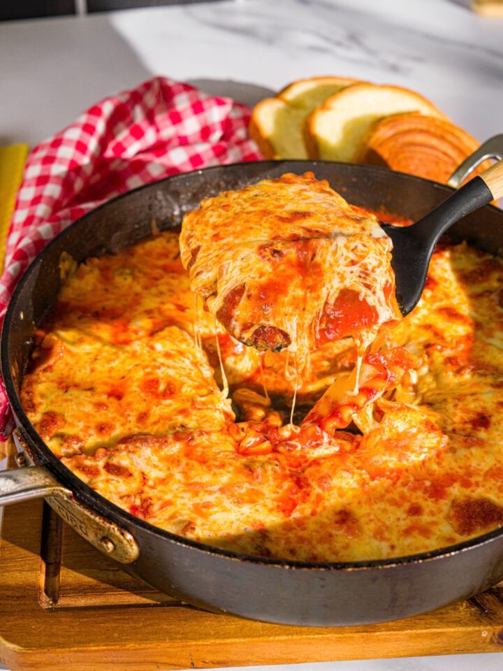 A spatula taking a slice of skillet lasagna from a skillet. The skillet is on a wooden board with a red checkered napkin.