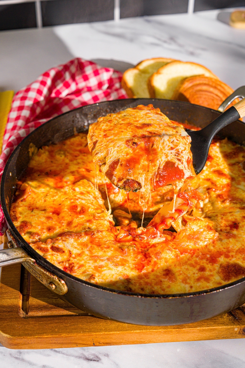A spatula taking a slice of skillet lasagna from a skillet. The skillet is on a wooden board with a red checkered napkin.