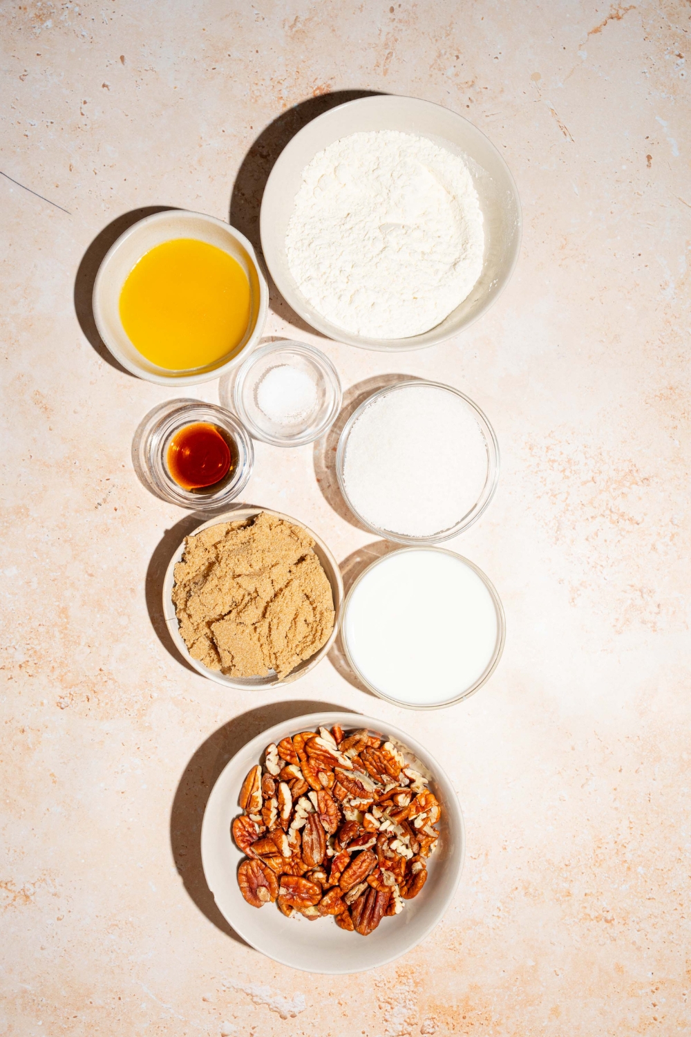 A tan counter with several bowls containing ingredients to make pecan pie cobbler including melted butter, milk, sugar, brown sugar, pecans, vanilla, and more.