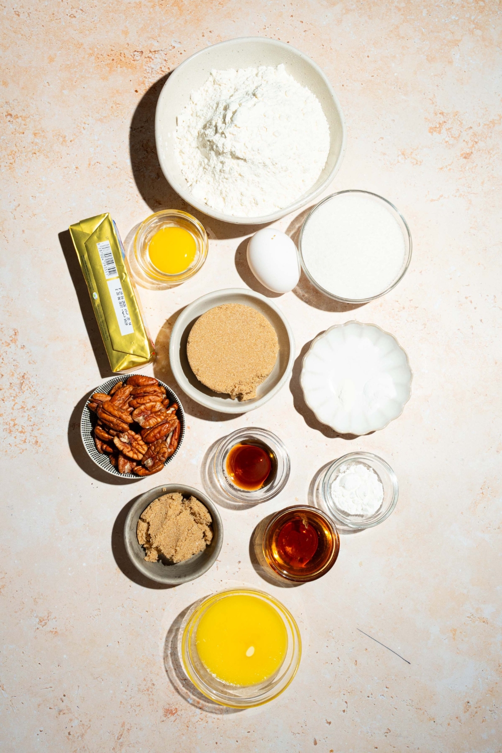 A tan counter with ingredients to make pecan pie cookies including flour, butter, egg, brown sugar, sugar, vanilla, pecans, and more.