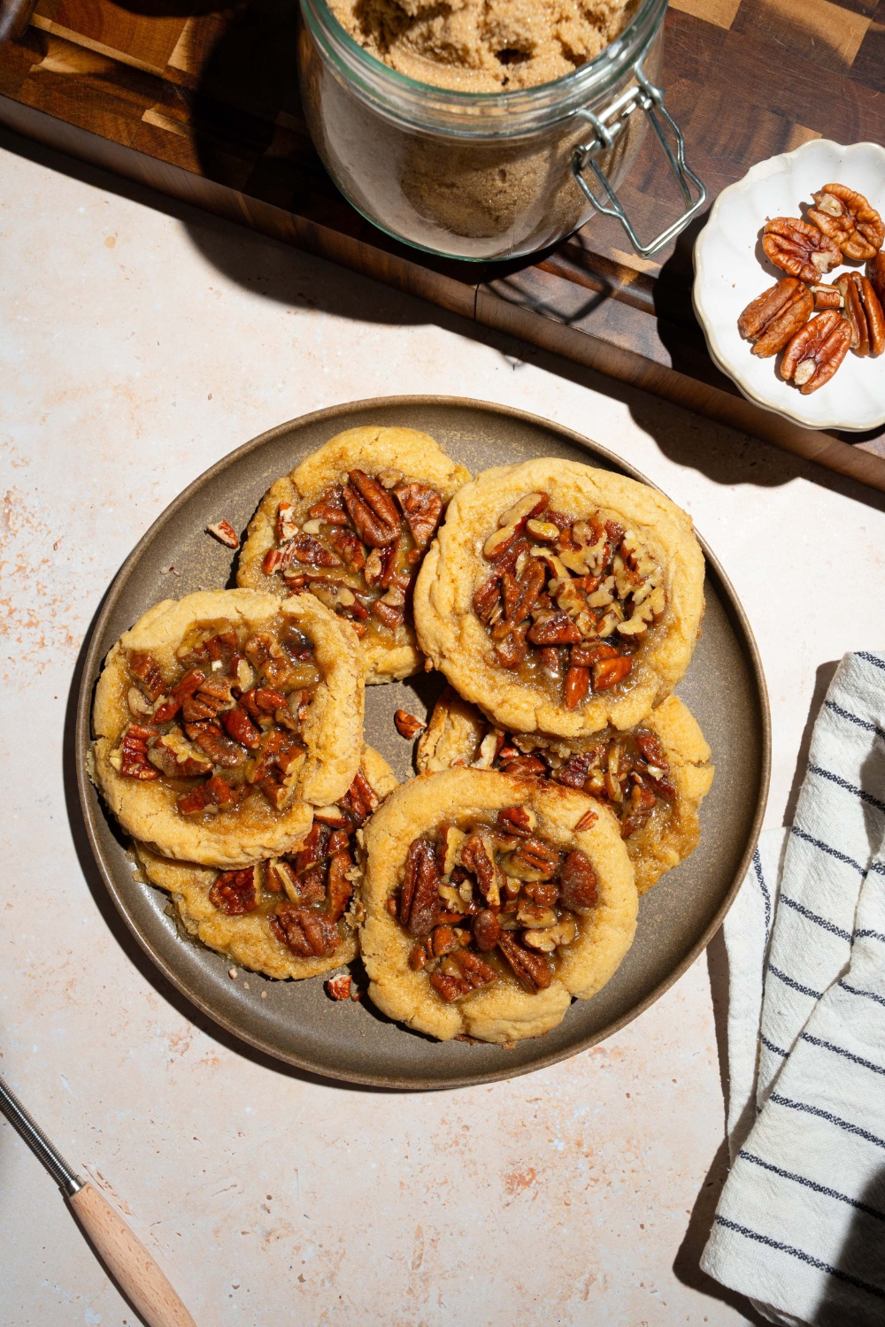 A plate with pecan pie cookies topped with pecan pie filling. The plate is on a tan counter with a white striped napkin and bowls of ingredients.