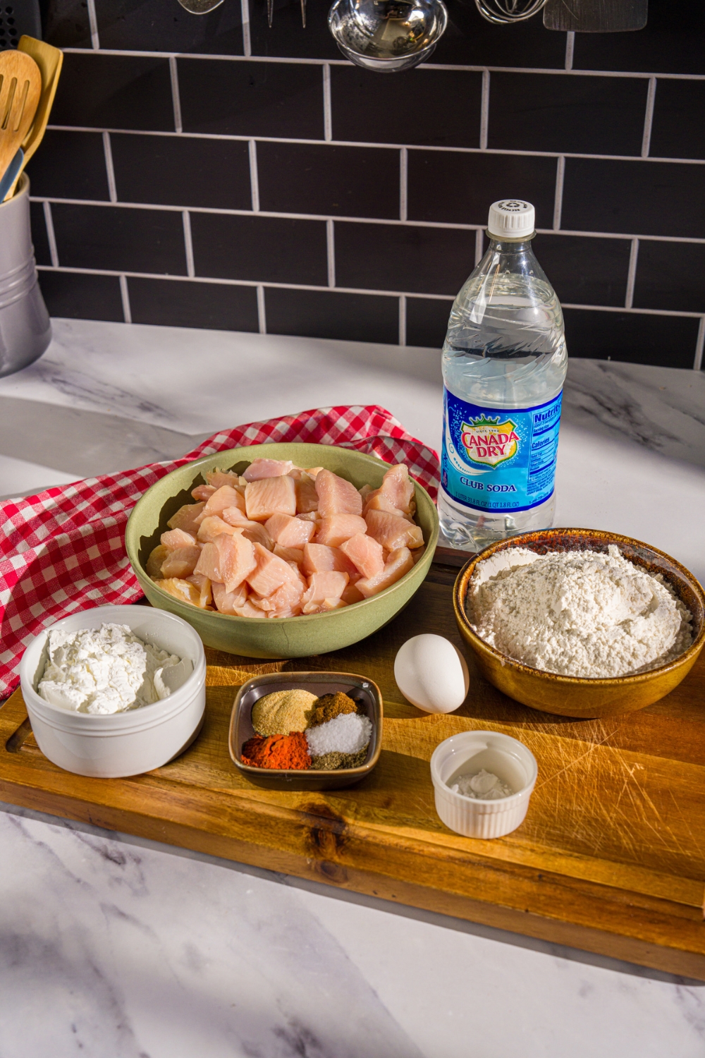 A wooden board with ingredients to make popcorn chicken including chicken pieces, flour, egg, club soda, cornstarch, and seasonings.