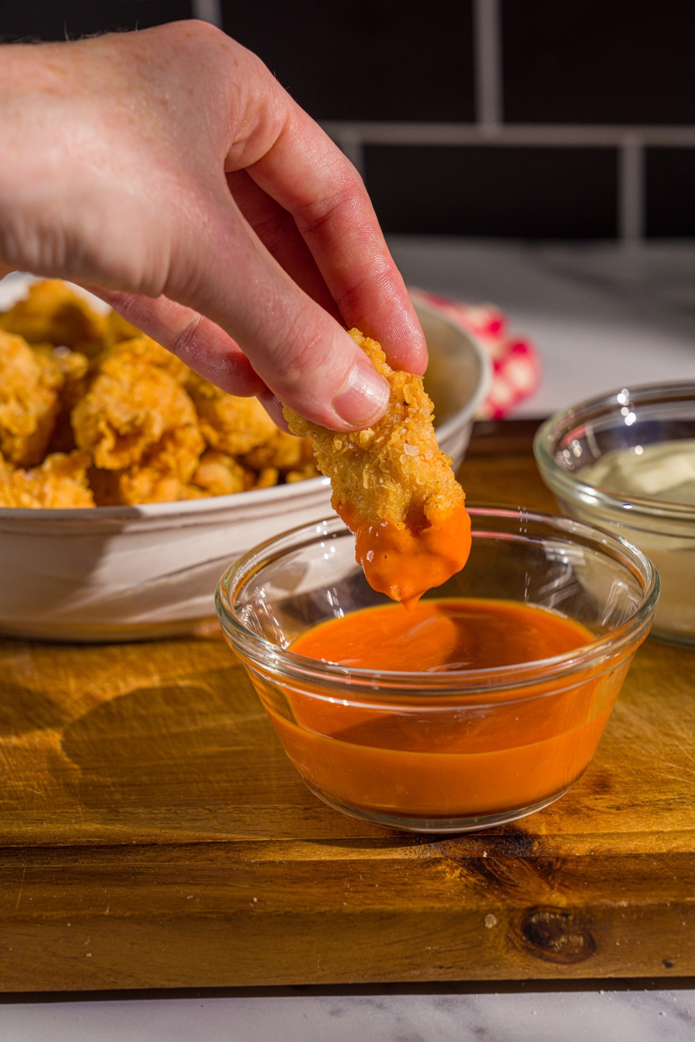 A hand dipping a piece of popcorn chicken into a small bowl of dipping sauce. The bowl is on a wooden board with a bowl of chicken.