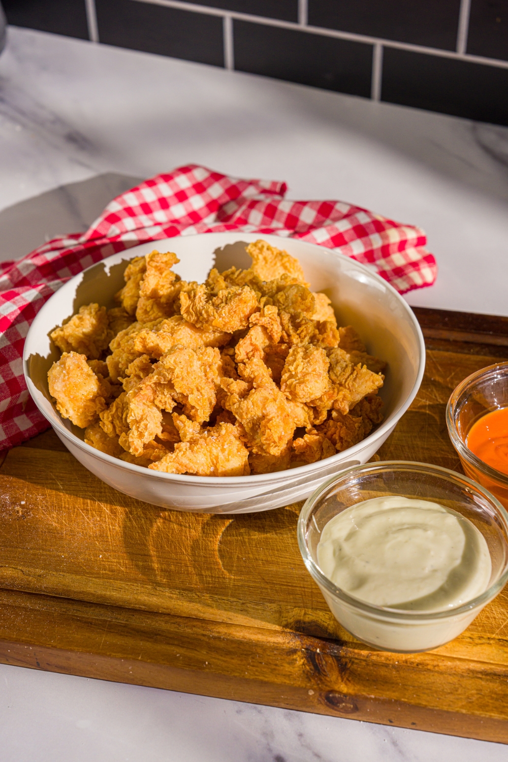 A bowl of popcorn chicken on a wooden board with two bowls of dipping sauces. The board is on a marble counter with a red striped napkin.