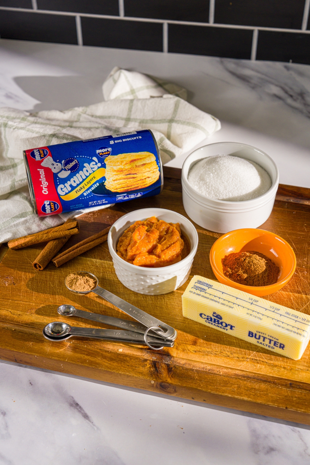 A wooden board with ingredients to make pumpkin pull apart bread including canned biscuits, pumpkin puree, butter, cinnamon, sugar, and pumpkin pie spice.