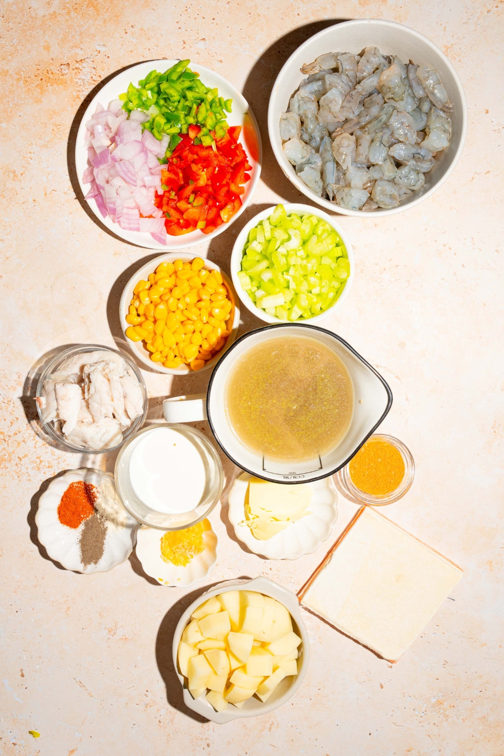 A tan counter with several bowls containing ingredients to make seafood pot pie including chicken broth, shrimp, crab meat, potatoes, puff pastry, celery, onion, bell peppers, butter, heavy cream, and more.