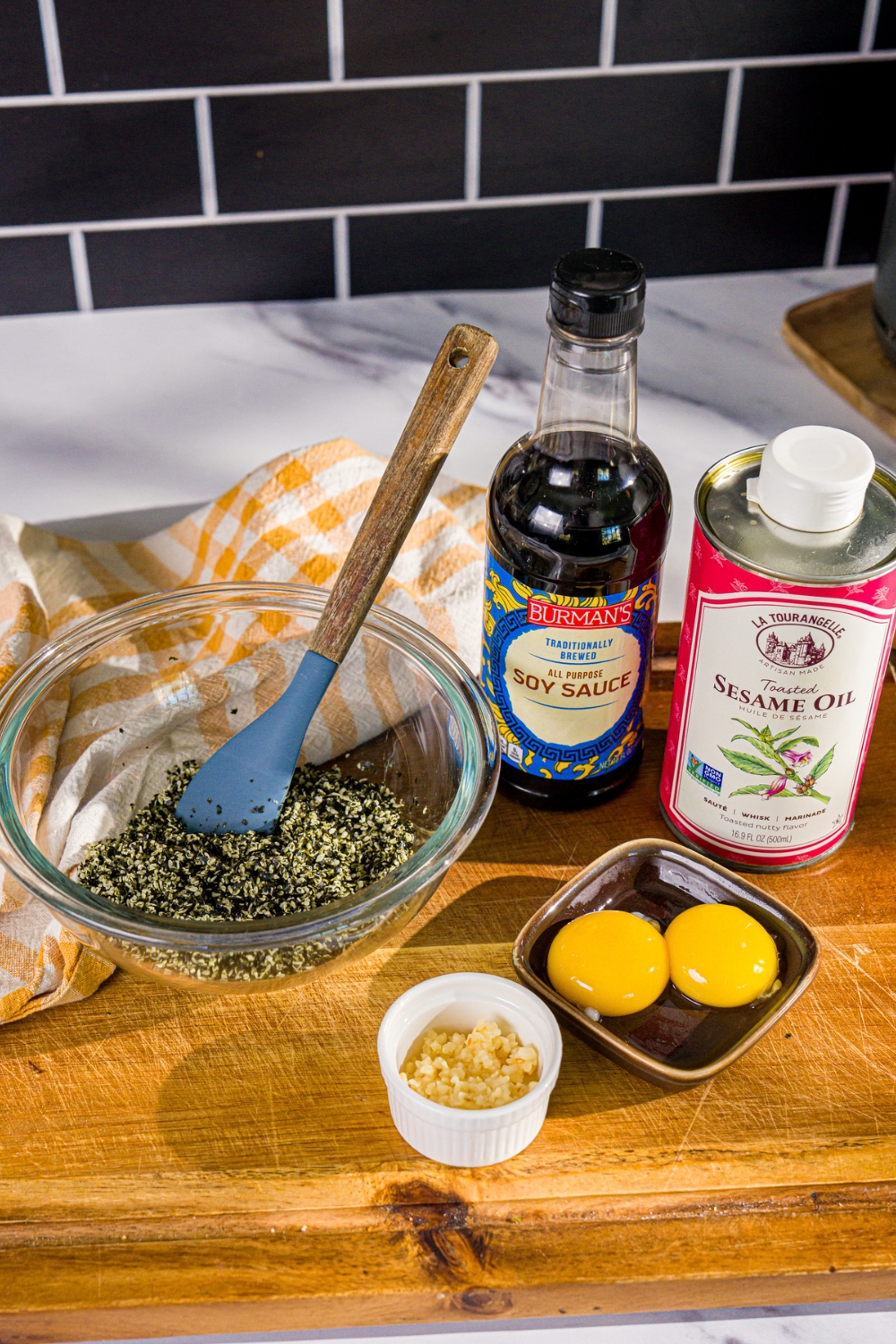 A wooden board with ingredients to make seaweed pasta including chopped seaweed and sesame seeds, garlic, egg yolk, sesame oil, and soy sauce.