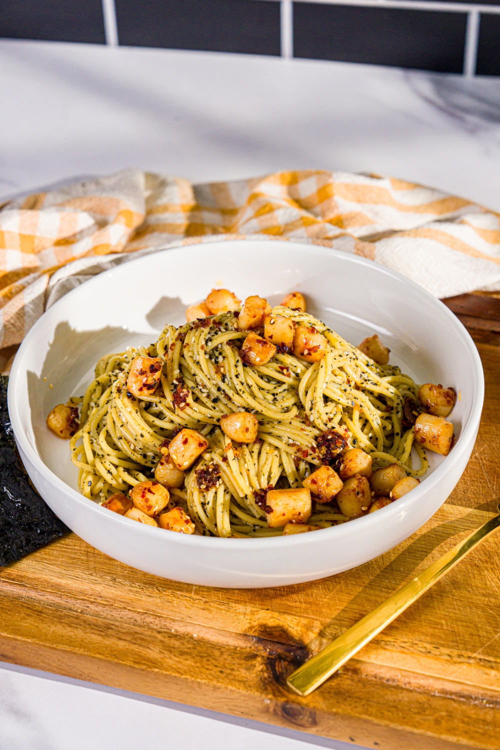 A bowl of seaweed pasta topped with seared scallops and chili crisp. The bowl is on a wooden board with a yellow cloth napkin.