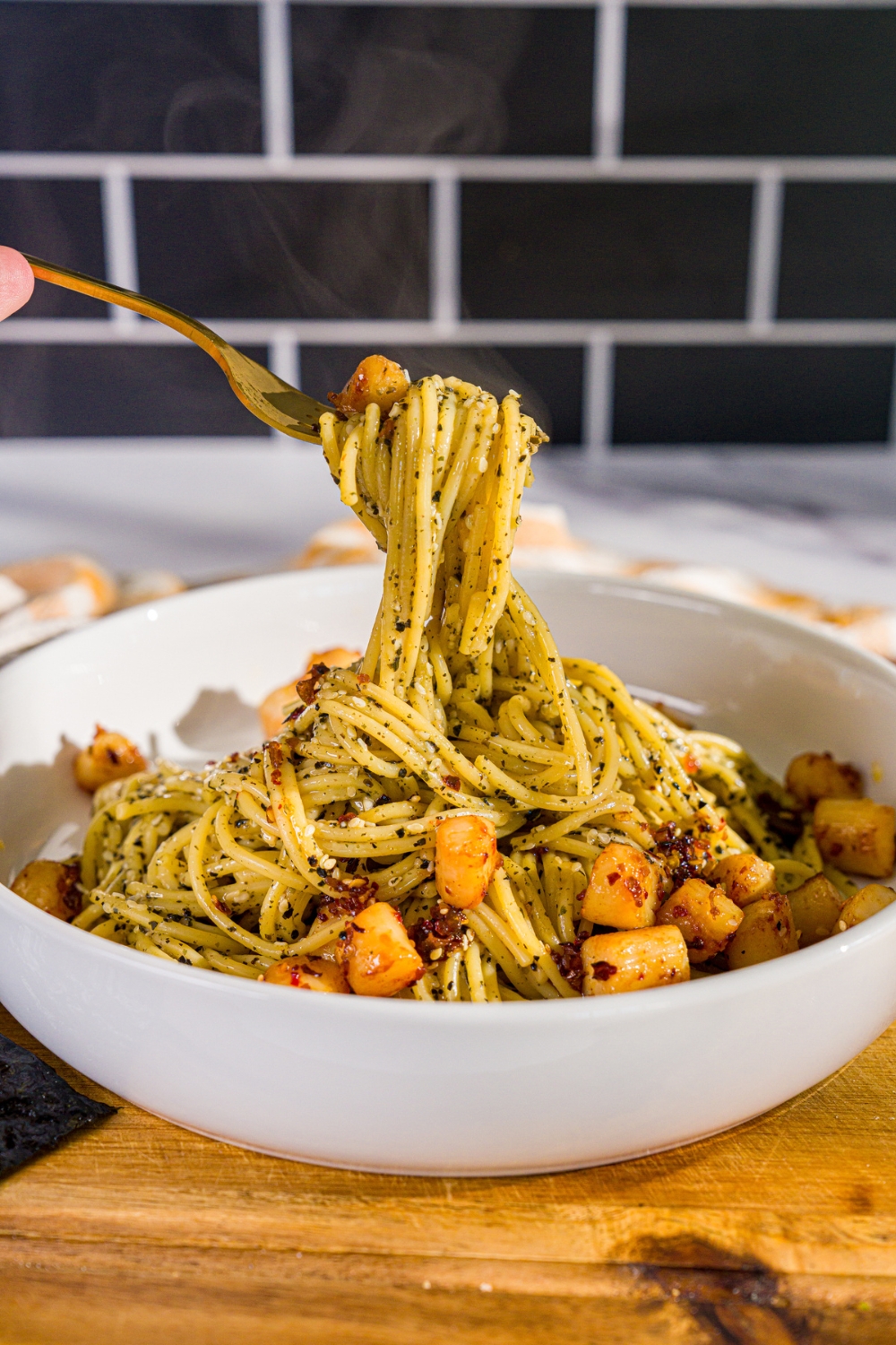 A bowl of seaweed pasta topped with seared scallops and chili crisp. A fork is pulling a bite from the bowl. The bowl is on a wooden board.
