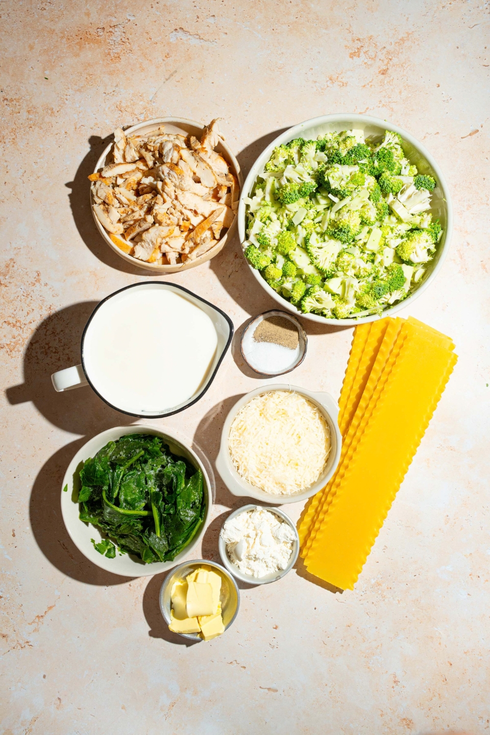 A tan counter with several bowls containing ingredients to make chicken broccoli lasagna including lasagna noodles, broccoli, garlic, chicken, milk, shredded cheese, flour, and more.