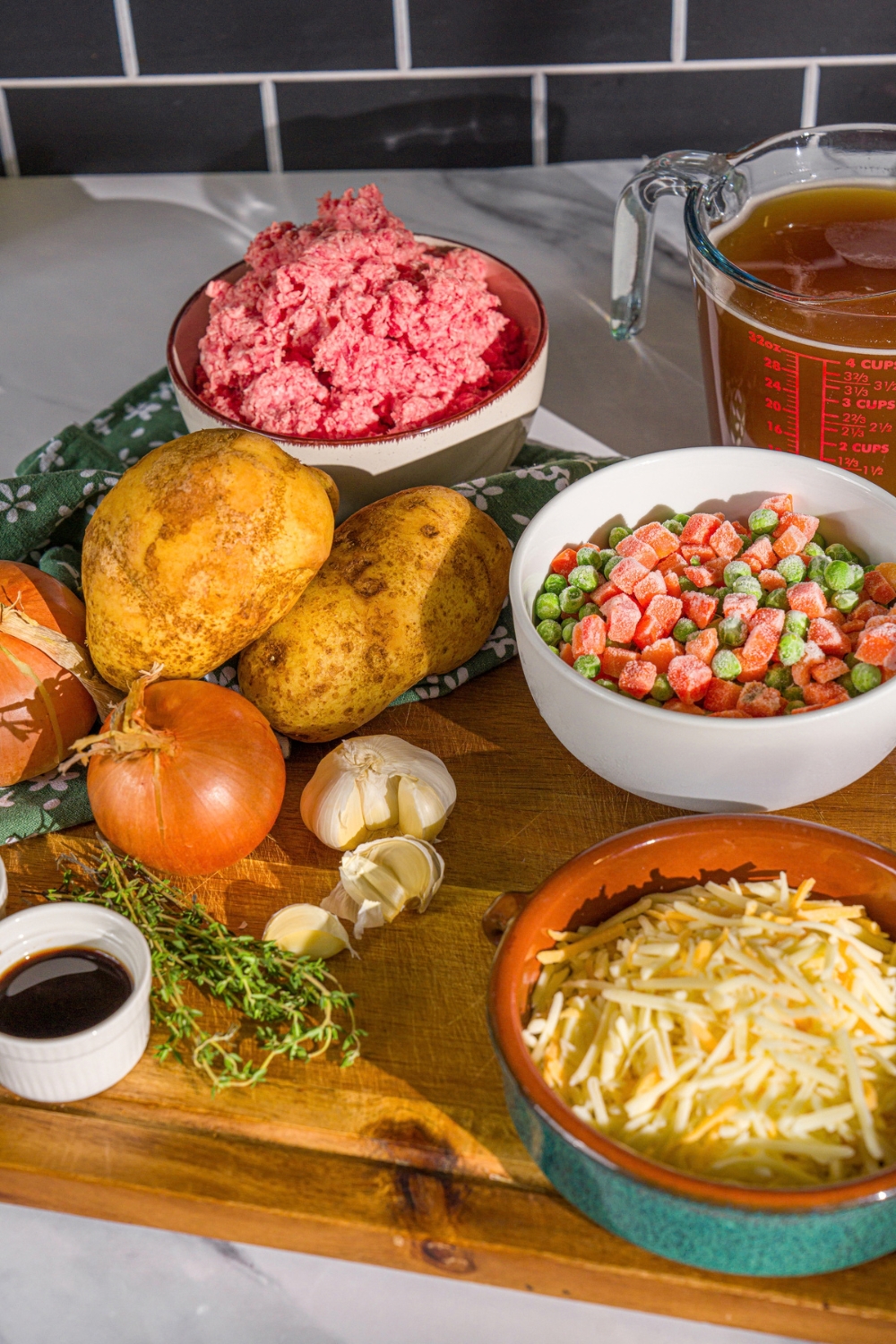 A wooden board with ingredients to make shepherd's pie soup including ground beef, frozen carrots and peas, onion, shredded cheese, beef stock, and potatoes.