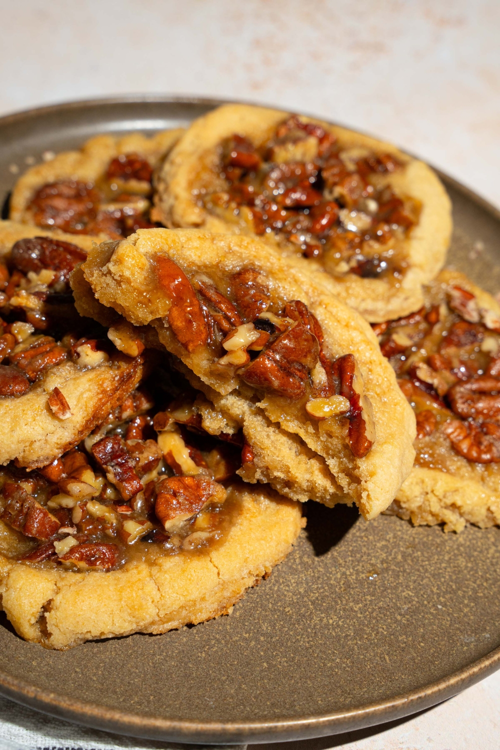 A plate with pecan pie cookies topped with pecan pie filling. One of the cookies is sliced in half. The plate is on a tan counter.