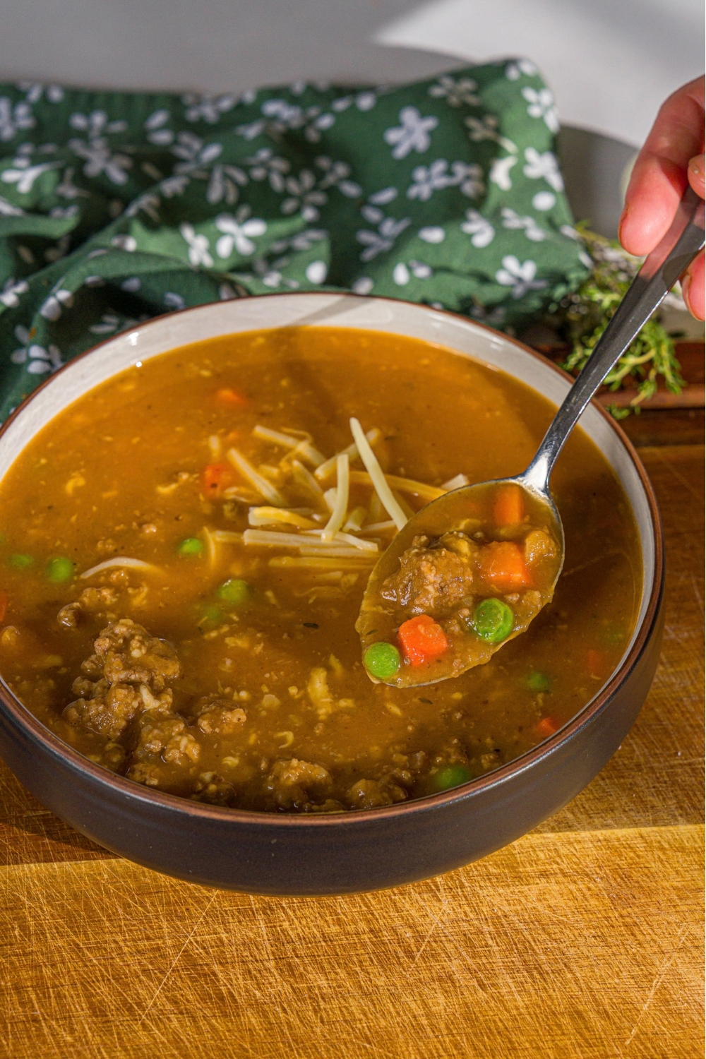 A bowl of shepherd's pie soup topped with shredded cheese. A spoon is taking a bite from the bowl. The bowl is on a wooden board with a green cloth napkin.