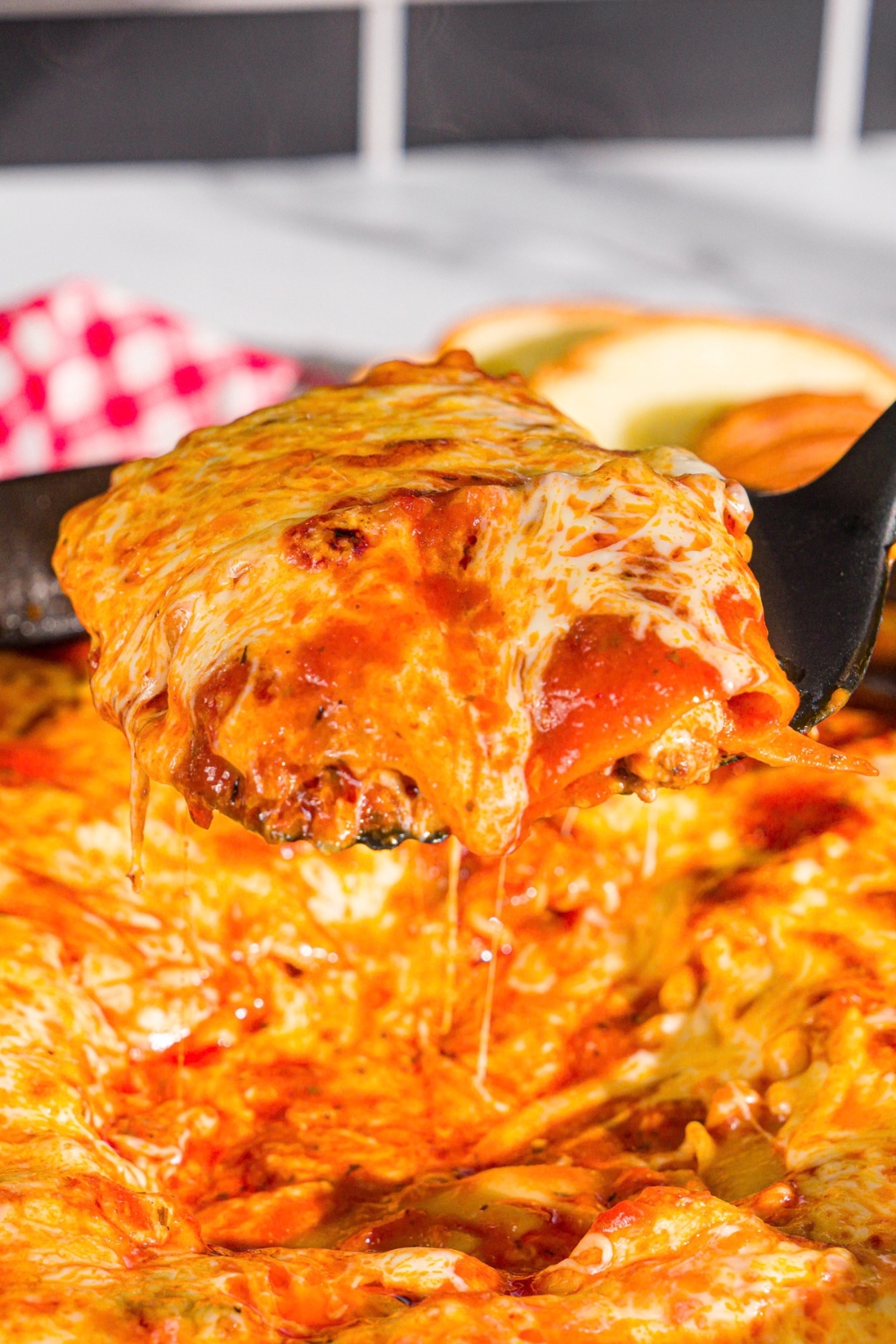 A spatula taking a slice of skillet lasagna from a skillet. The skillet is on a wooden board with a red checkered napkin.