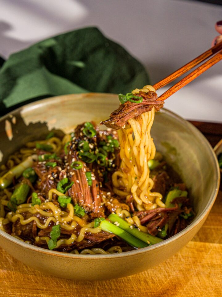 A bowl of crockpot Mongolian beef served with noodles and garnished with sliced green onions and sesame seeds. A pair of chopsticks are pulling a bite from the bowl. The bowl is on a wooden board with a green cloth napkin.