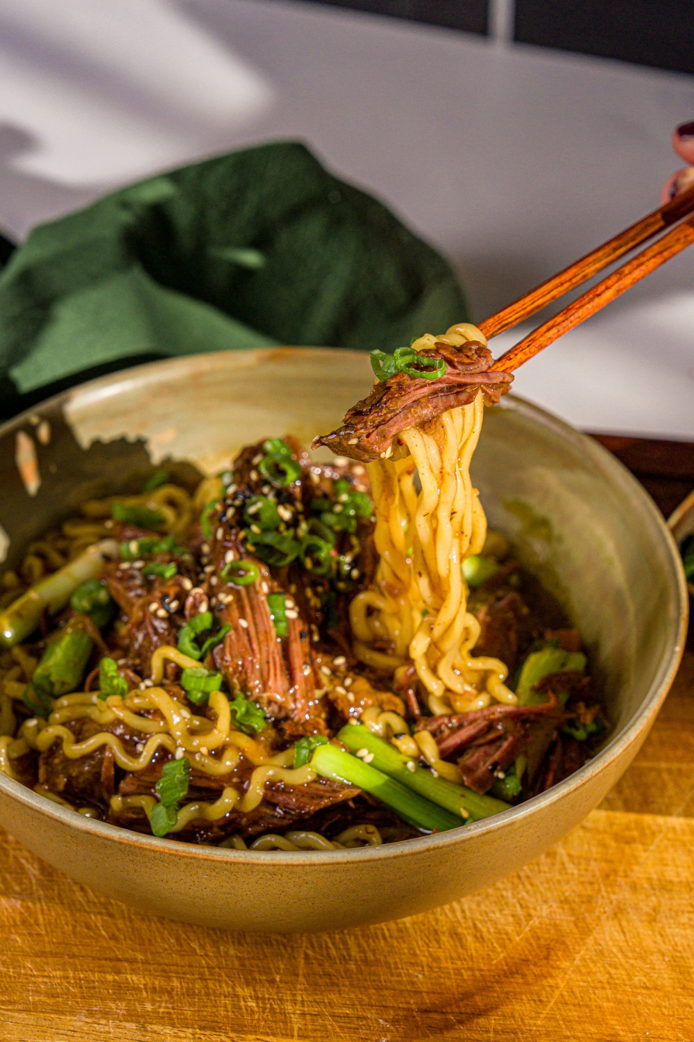 A bowl of crockpot Mongolian beef served with noodles and garnished with sliced green onions and sesame seeds. A pair of chopsticks are pulling a bite from the bowl. The bowl is on a wooden board with a green cloth napkin.