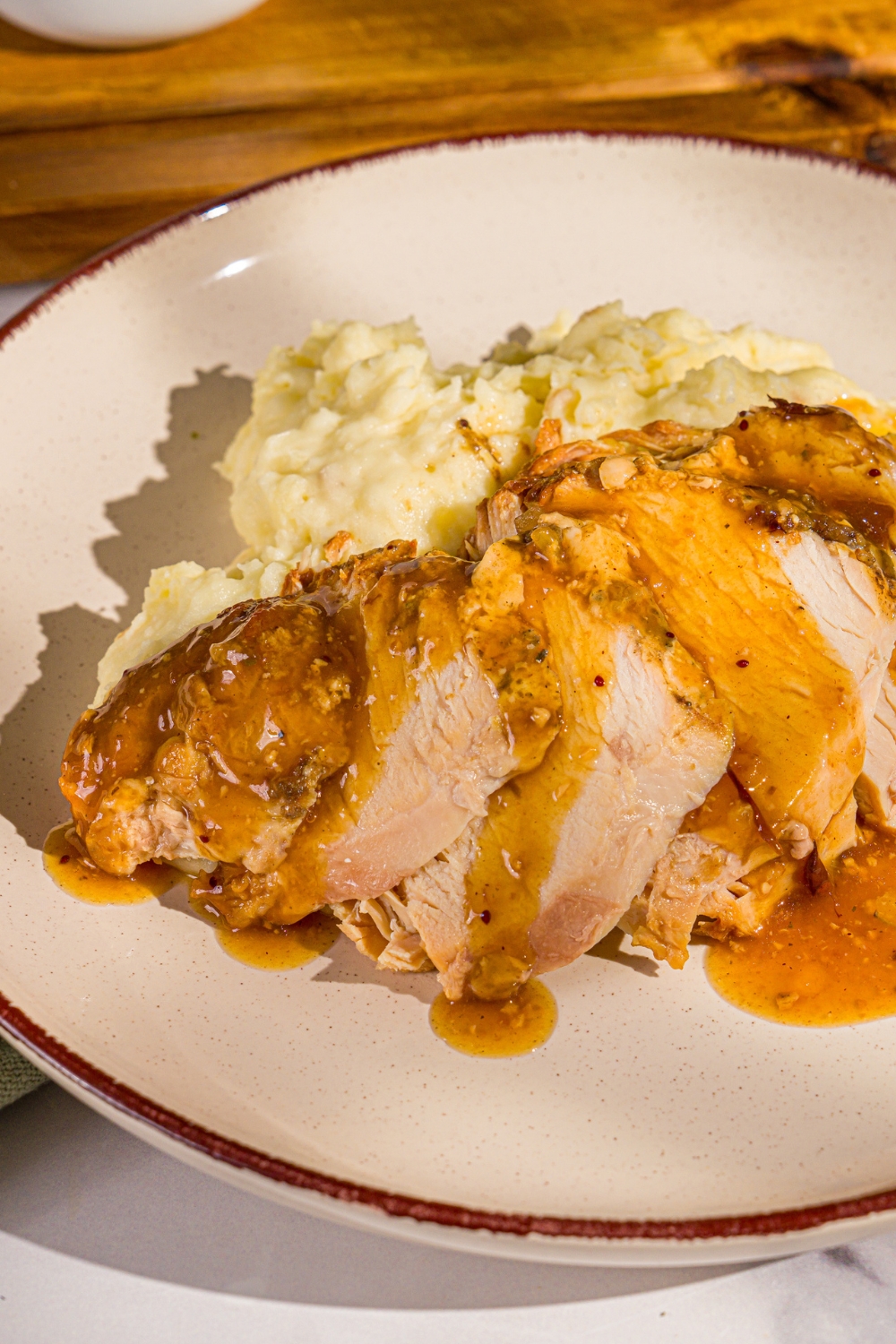A white plate with sliced crock pot butterball turkey breast topped with gravy and served with mashed potatoes. The plate is on a marble counter with a wooden board.
