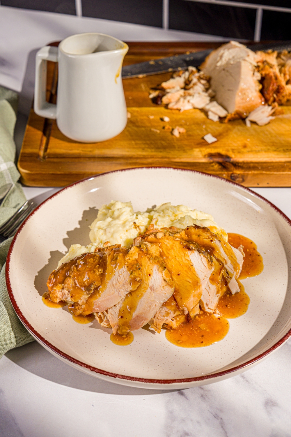 A white plate with sliced crock pot butterball turkey breast topped with gravy and served with mashed potatoes. The plate is on a marble counter with a wooden board with turkey breast.