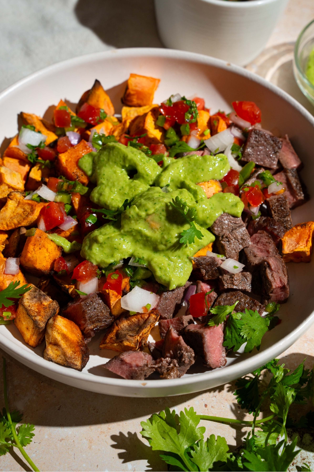 A sweet potato steak bowl topped with pico de gallo and avocado cilantro lime dressing. The bowl is on a tan counter with fresh cilantro.