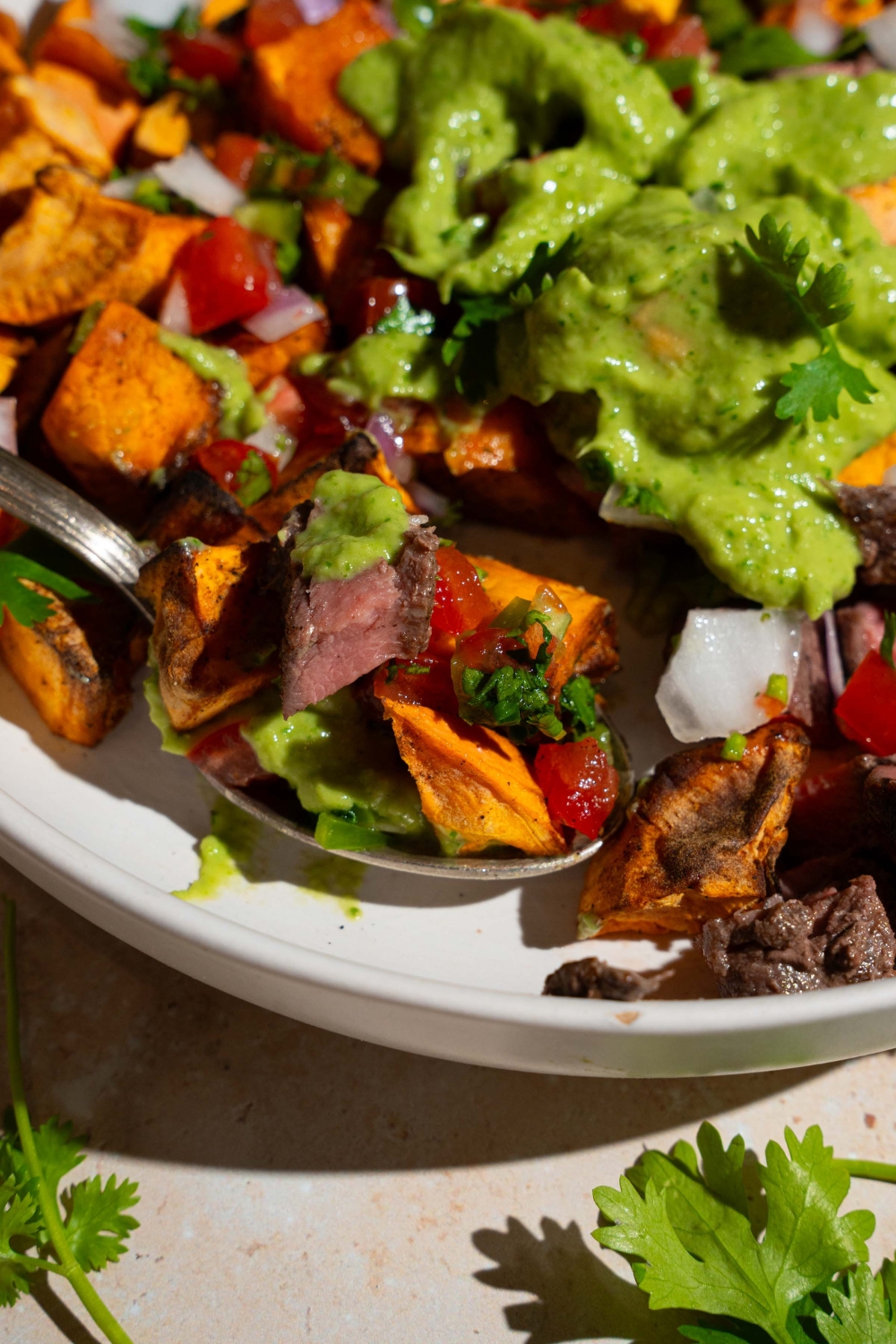A sweet potato steak bowl topped with pico de gallo and avocado cilantro lime dressing. A spoon is taking a bite from the bowl. The bowl is on a tan counter with fresh cilantro.
