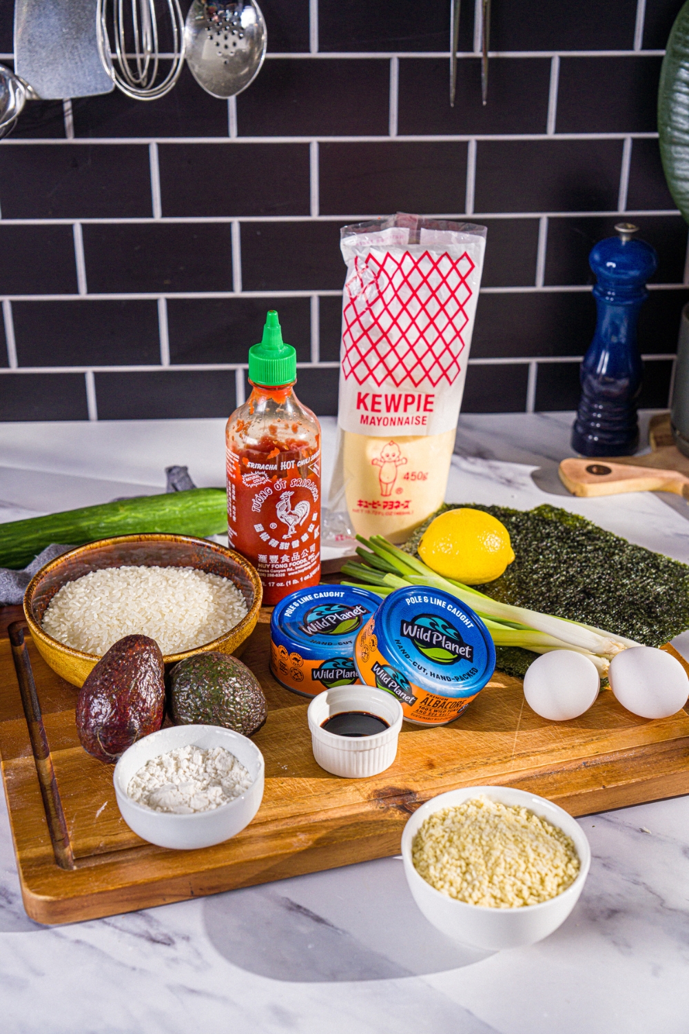 A wooden board with ingredients to make sushi burrito including seaweed wraps, sushi rice. tuna, avocado, cucumber, lemon, eggs, panko breading, kewpie mayo, and sriracha.