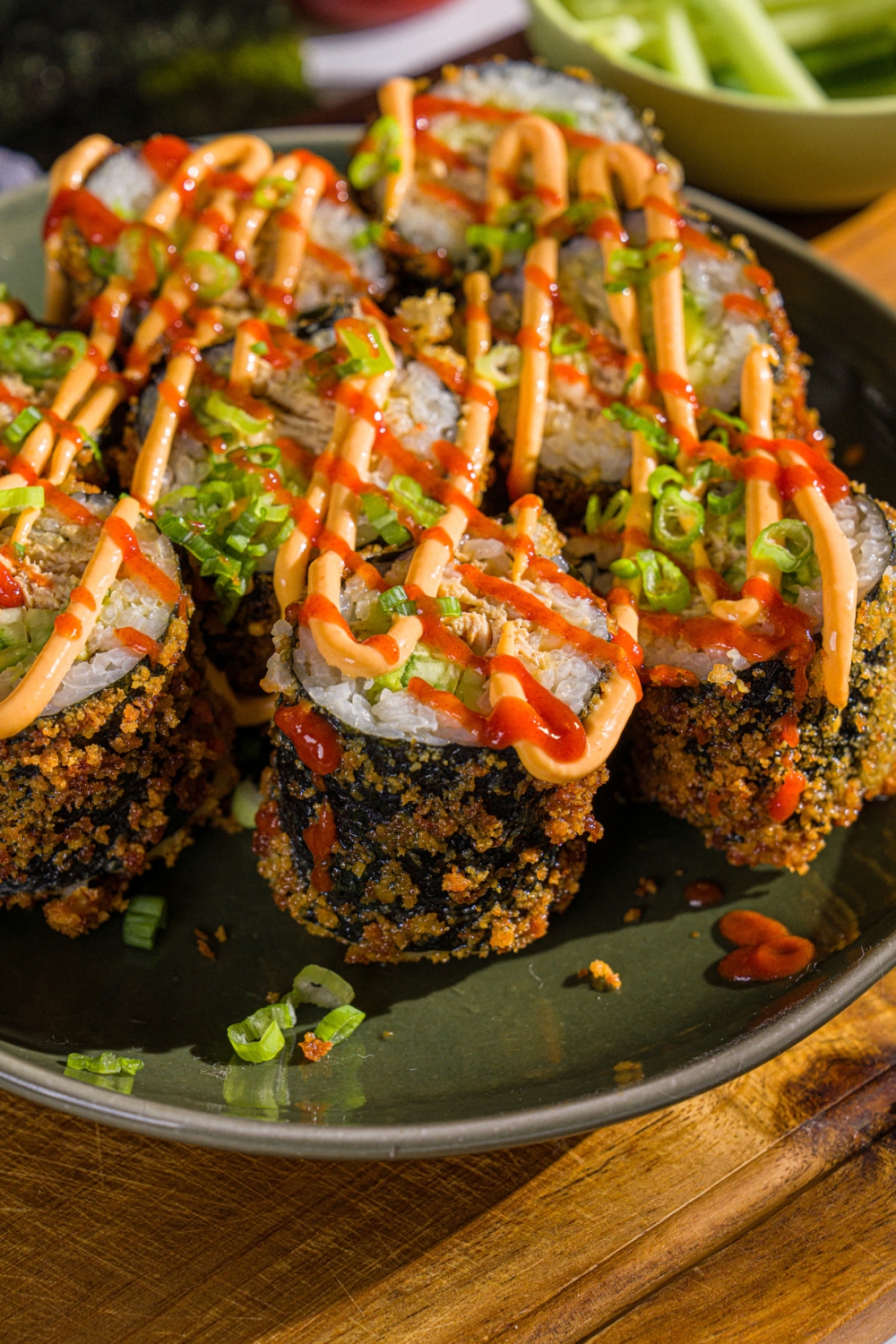 A plate of sliced fried sushi burrito topped with spicy mayo, sriracha, and sliced green onions. The plate is on a wooden board.