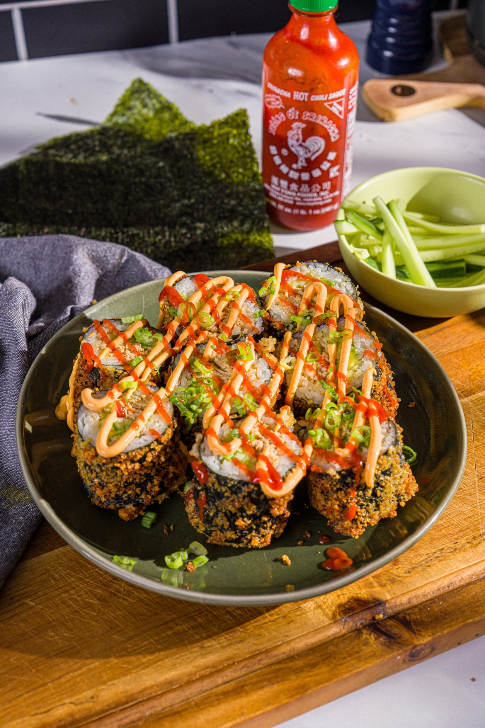 A plate of sliced fried sushi burrito topped with spicy mayo, sriracha, and sliced green onions. The plate is on a wooden board with a bowl of sliced cucumber and seaweed wraps.