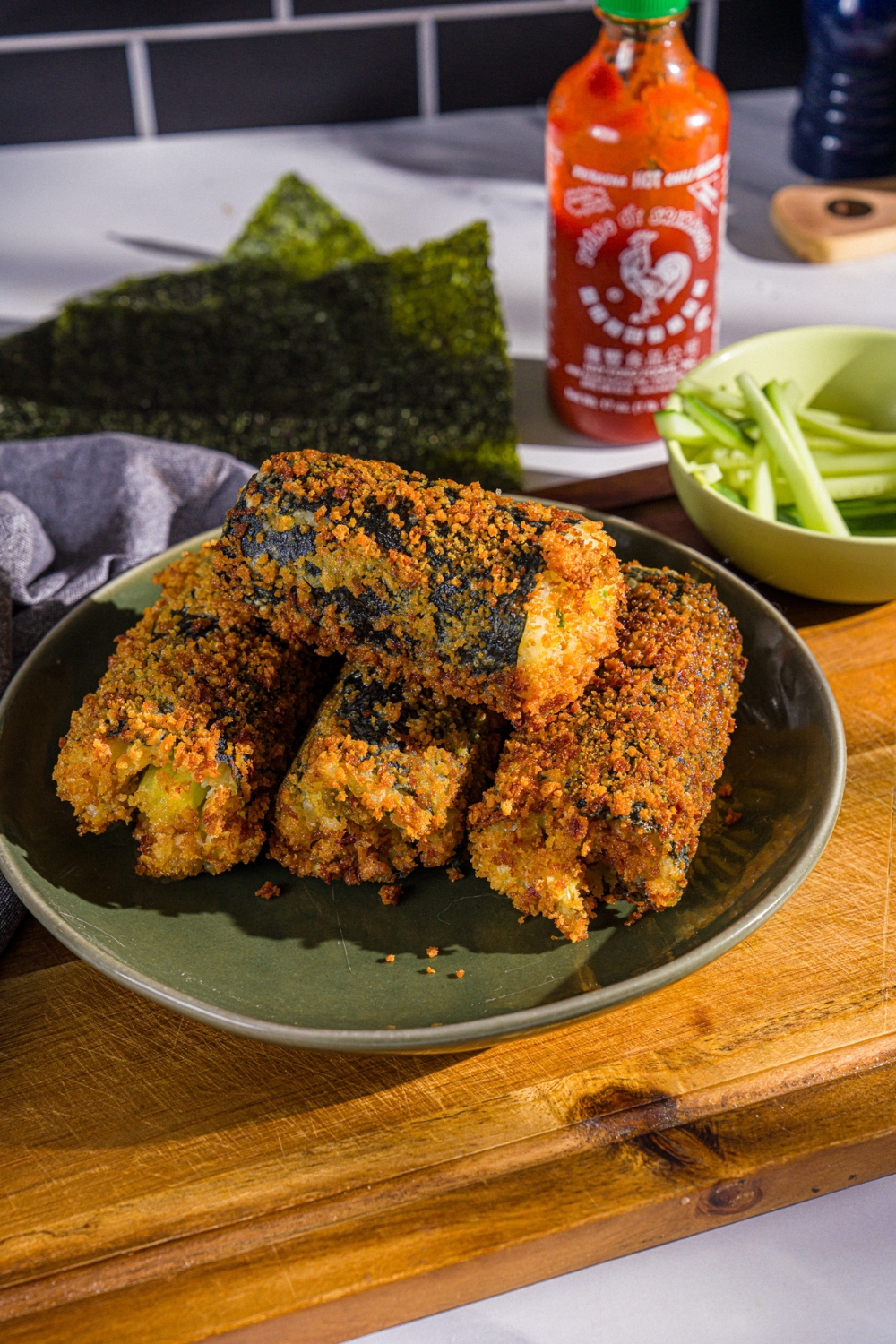 A plate with fried sushi burrito pieces. The plate is on a wooden board with a bowl of sliced cucumber, seaweed wraps, and sriracha.