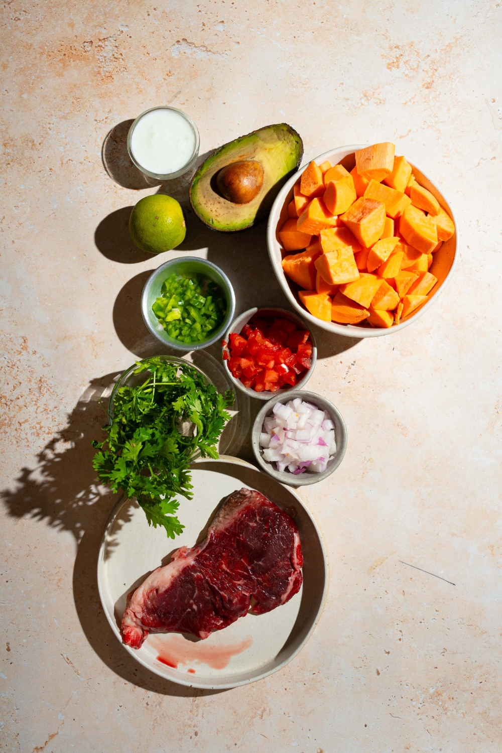 A tan counter with ingredients to make sweet potato steak bowl including sweet potatoes, steak, avocado, cilantro, tomatoes, onion, lime, and more.
