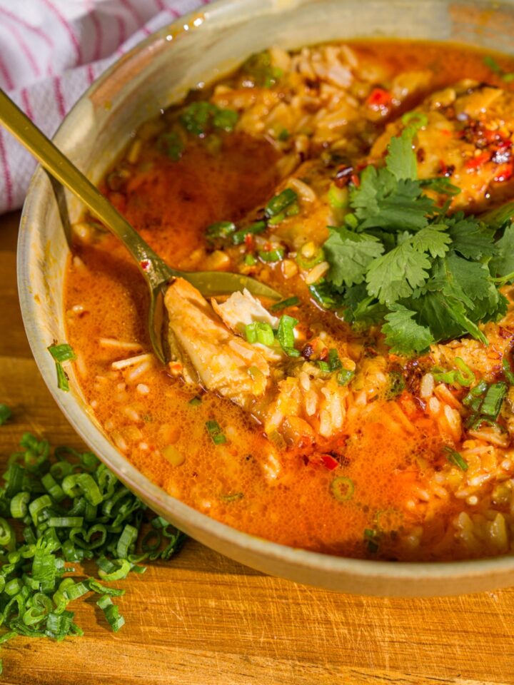 A bowl of thai coconut chicken bowl garnished with sliced green onion, cilantro, and chili oil. A spoon is dipped in the bowl. The bowl is on a wooden board.