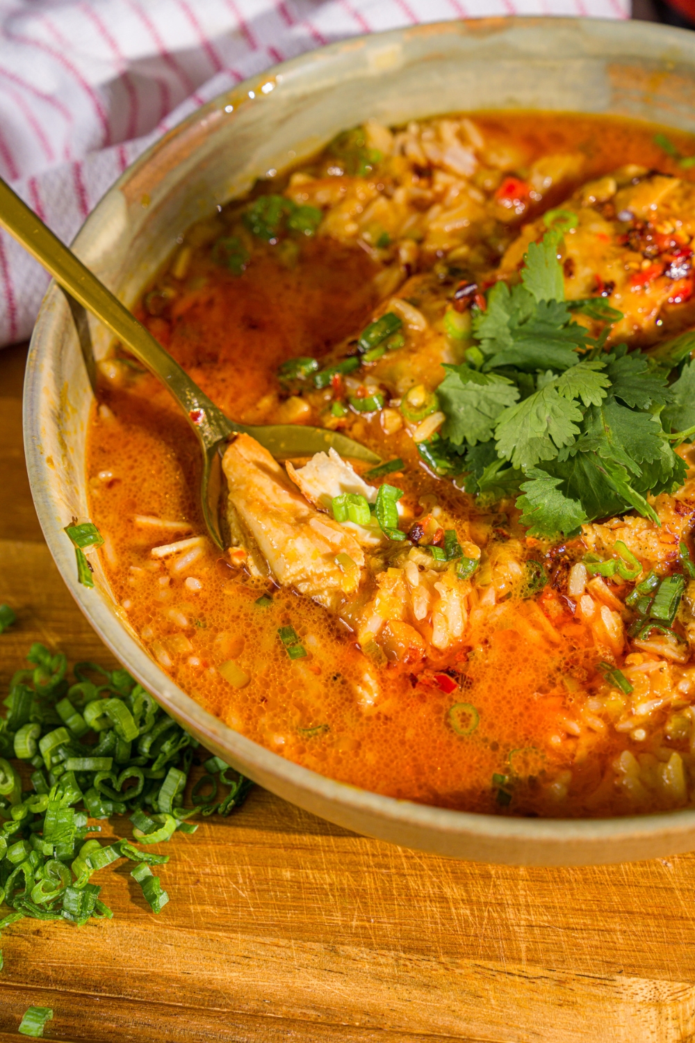 A bowl of thai coconut chicken bowl garnished with sliced green onion, cilantro, and chili oil. A spoon is dipped in the bowl. The bowl is on a wooden board.
