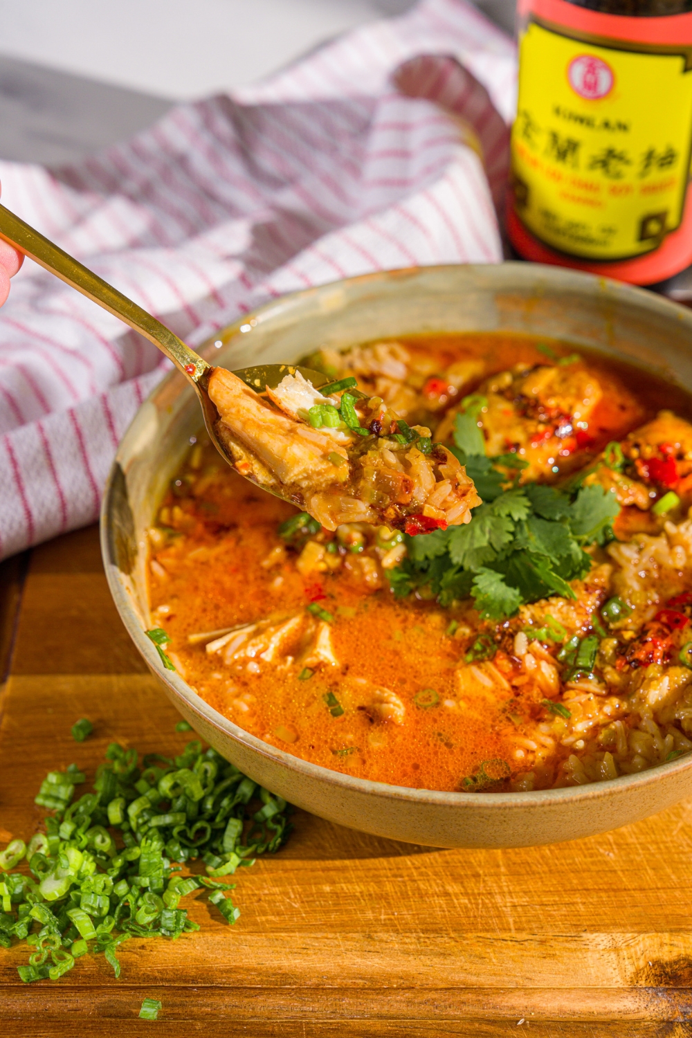 A bowl of thai coconut chicken bowl garnished with sliced green onion, cilantro, and chili oil. A spoon is dipped in the bowl. The bowl is on a wooden board with sliced green onion and a white cloth napkin.