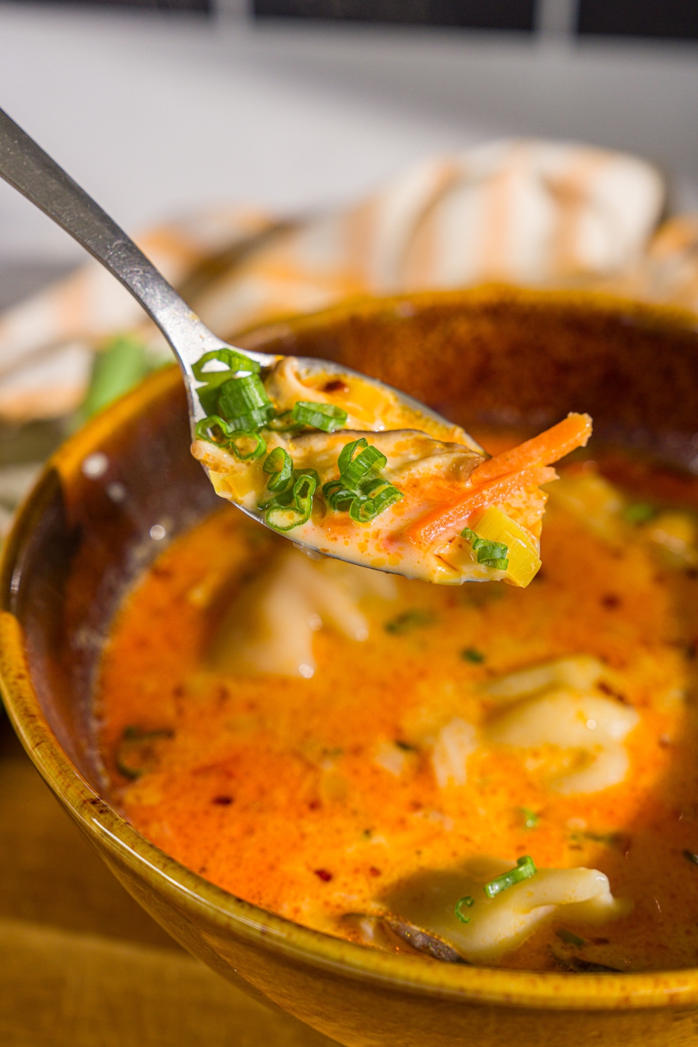 A close up of a spoon with a bite of creamy potsticker soup garnished with sliced green onion and chili crisp. There. is a bowl of soup blurred in the background.