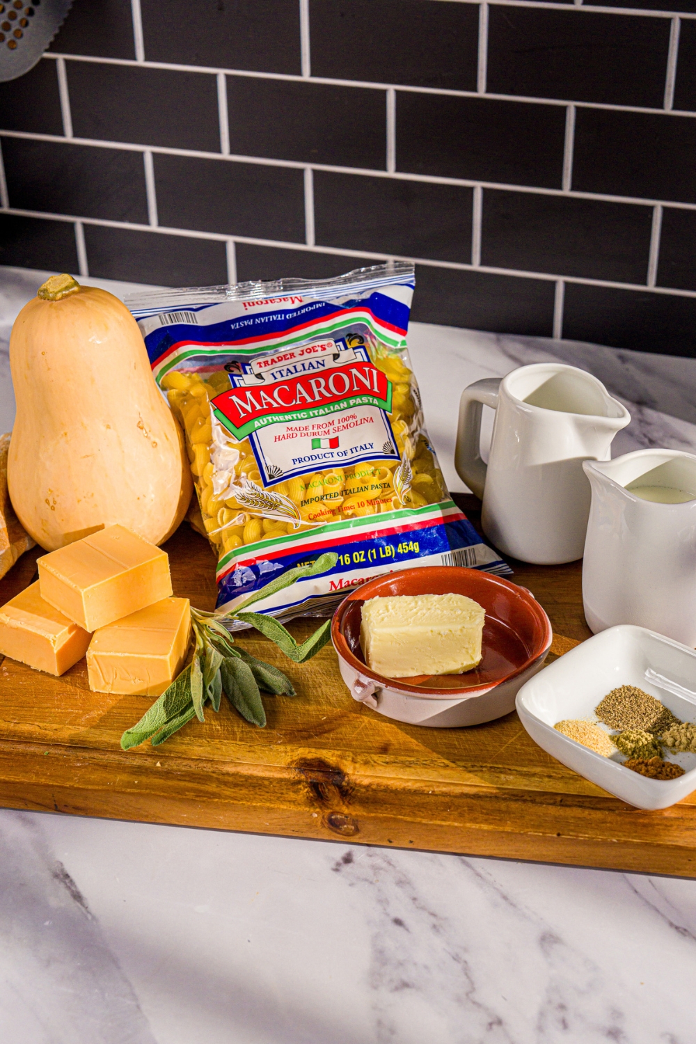 A package of Trader Joe's Italian macaroni, a block of cheddar cheese, a whole butternut squash, a pitcher of milk, a pitcher of heavy cream, butter, and a bowl of sage, garlic powder, nutmeg, and black pepper.