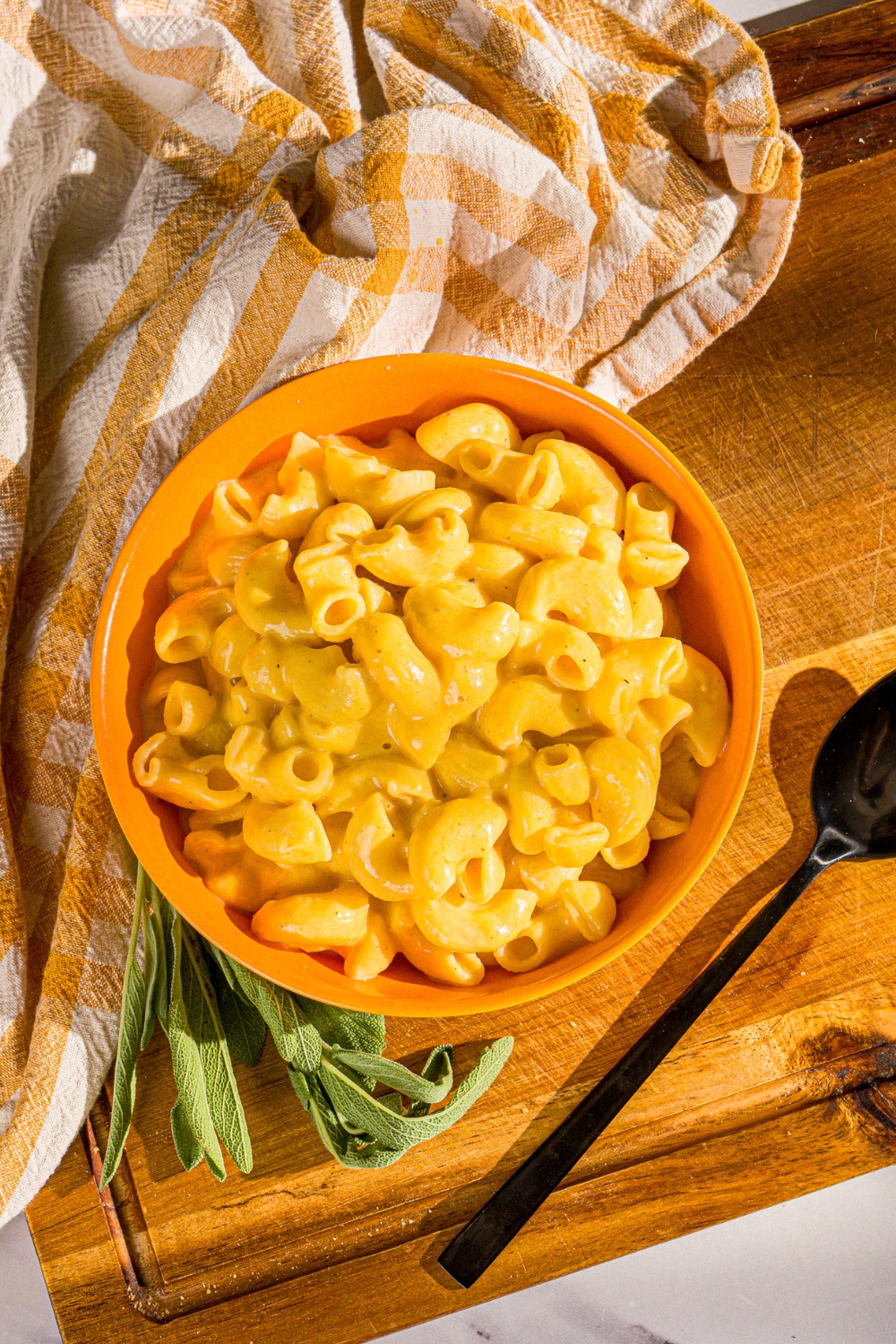 Butternut squash mac and cheese in an orange bowl on a wood board with a yellow and white stripped towel on the edge of the left side of the bowl.