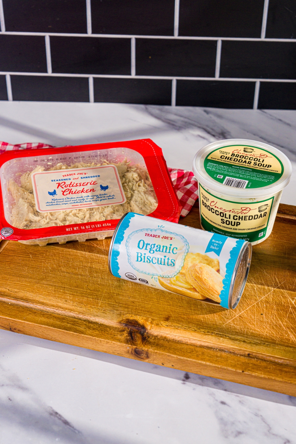 A wooden board with ingredients to make Trader Joe's chicken pot pie including Trader Joe's shredded rotisserie chicken, Trader Joe's jumbo biscuits, and Trad Joe's broccoli cheddar soup.