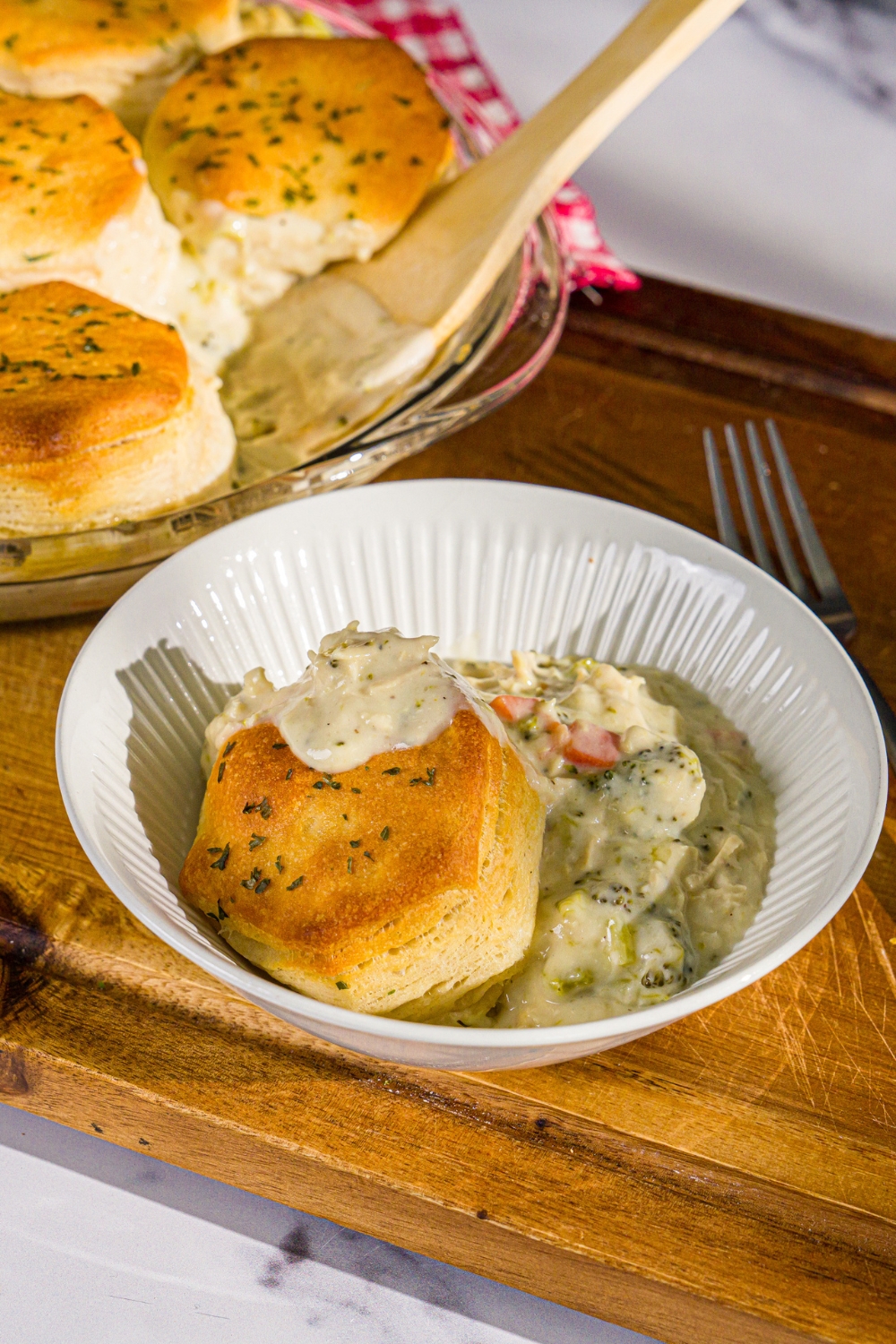 A bowl of Trader Joe's chicken pot pie with a biscuit topping. The bowl is on a wooden board with a fork and dish of pot pie.