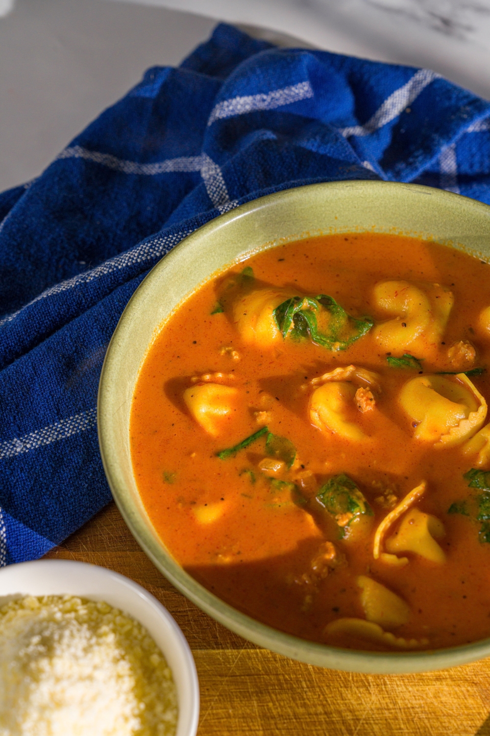 A bowl of creamy tomato tortellini soup. The bowl is on a wooden board with a small bowl of cheese and a blue cloth napkin.