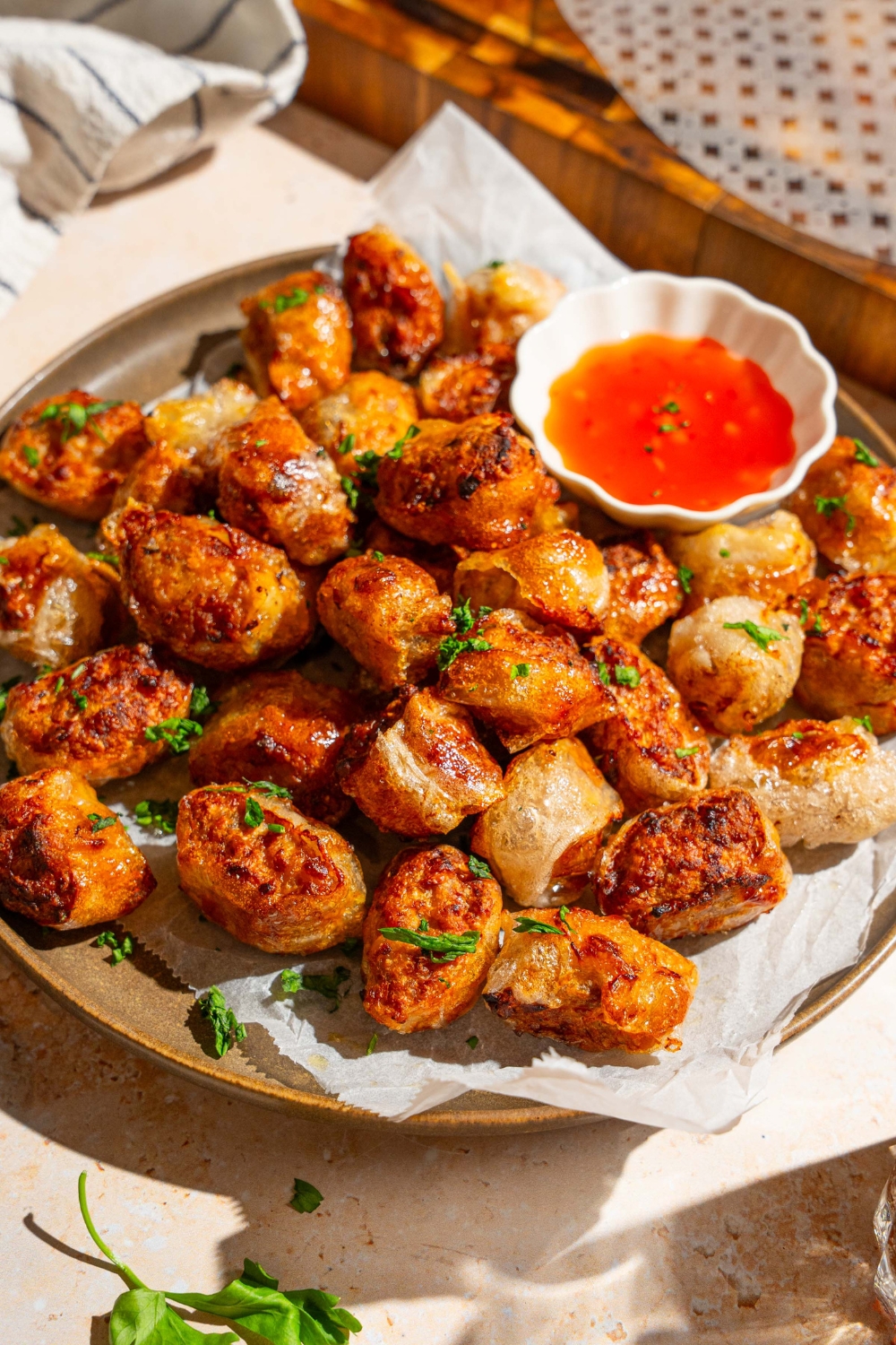 A plate lined with parchment paper with crispy rice paper chicken bites garnished with fresh parsley. There is a bowl of dipping sauce on the plate. The plate is on a tan counter.