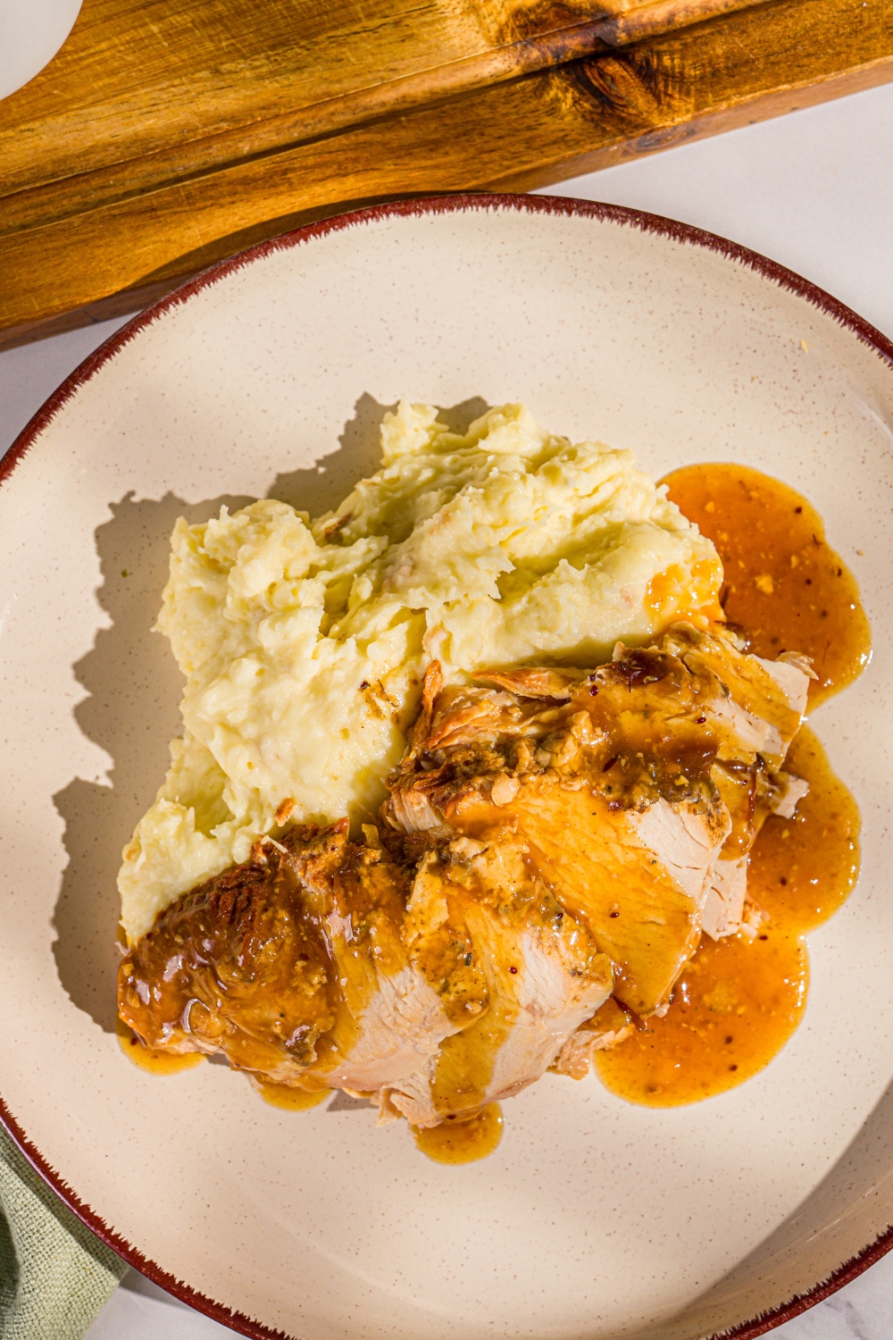 A white plate with sliced crock pot butterball turkey breast topped with gravy and served with mashed potatoes. The plate is on a marble counter with a wooden board.