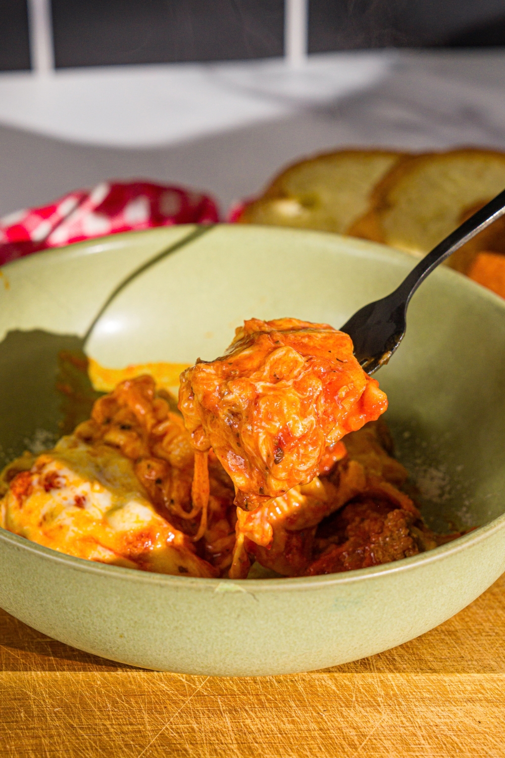 A bowl with a slice of skillet lasagna. A fork is taking a bite from the bowl. The bowl is on a wooden board with a red checkered napkin.