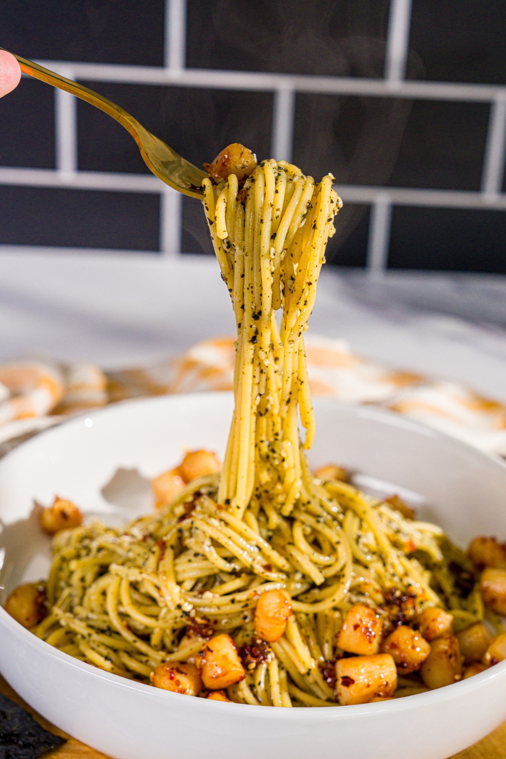 A bowl of seaweed pasta topped with seared scallops and chili crisp. A fork is pulling a bite from the bowl. The bowl is on a wooden board.