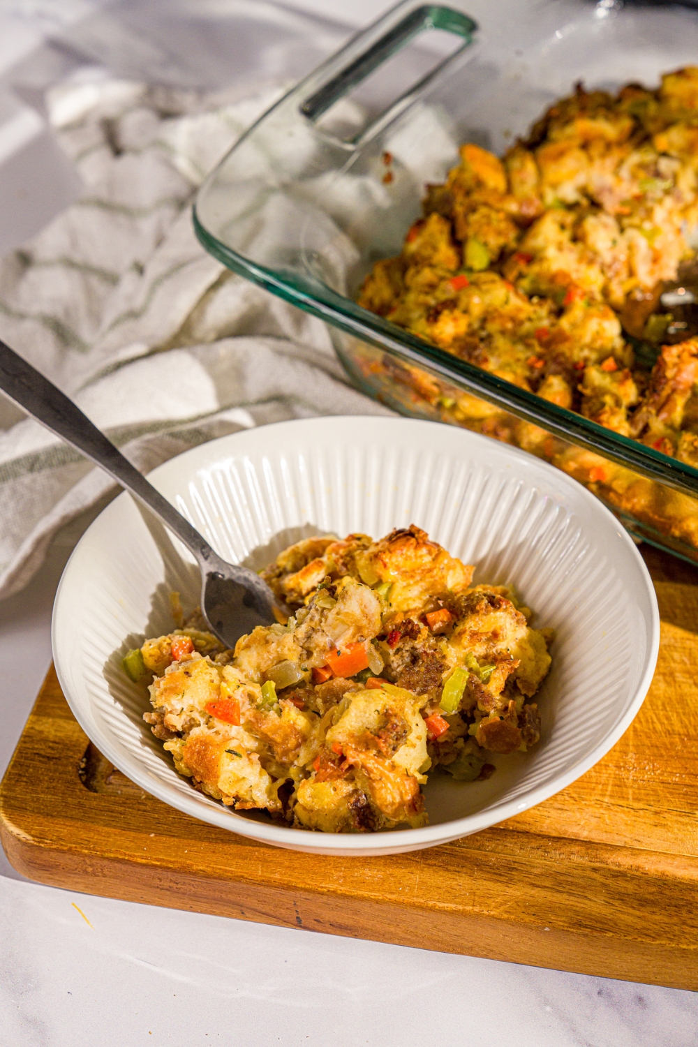 A bowl of White Castle stuffing with a fork in the bowl. The bowl is on a wooden board with a baking dish of stuffing.