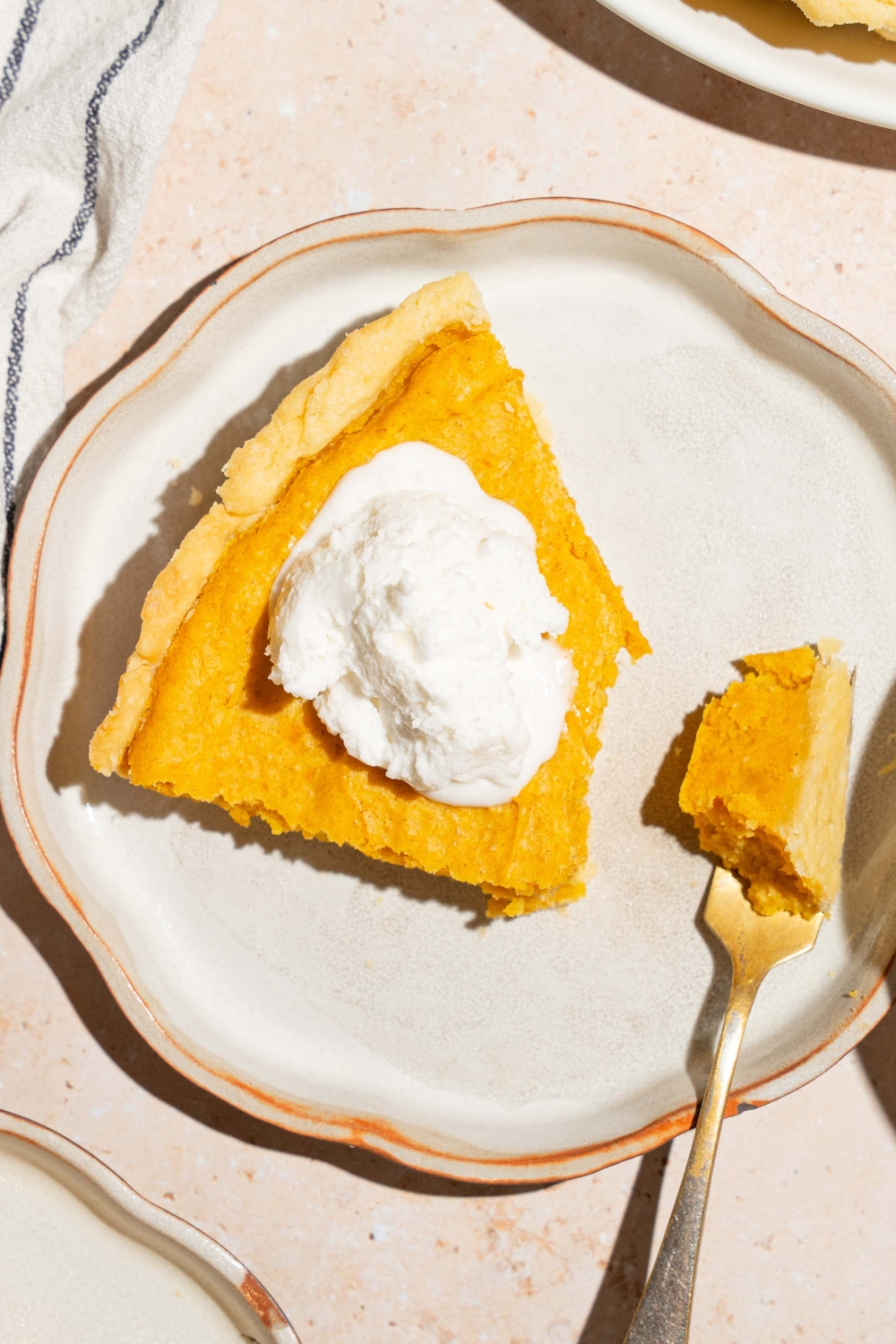 A plate with a slice of cream cheese pumpkin pie topped with a dollop of whipped cream. A fork is on the plate with a bite of pie. The plate is on a tan counter.